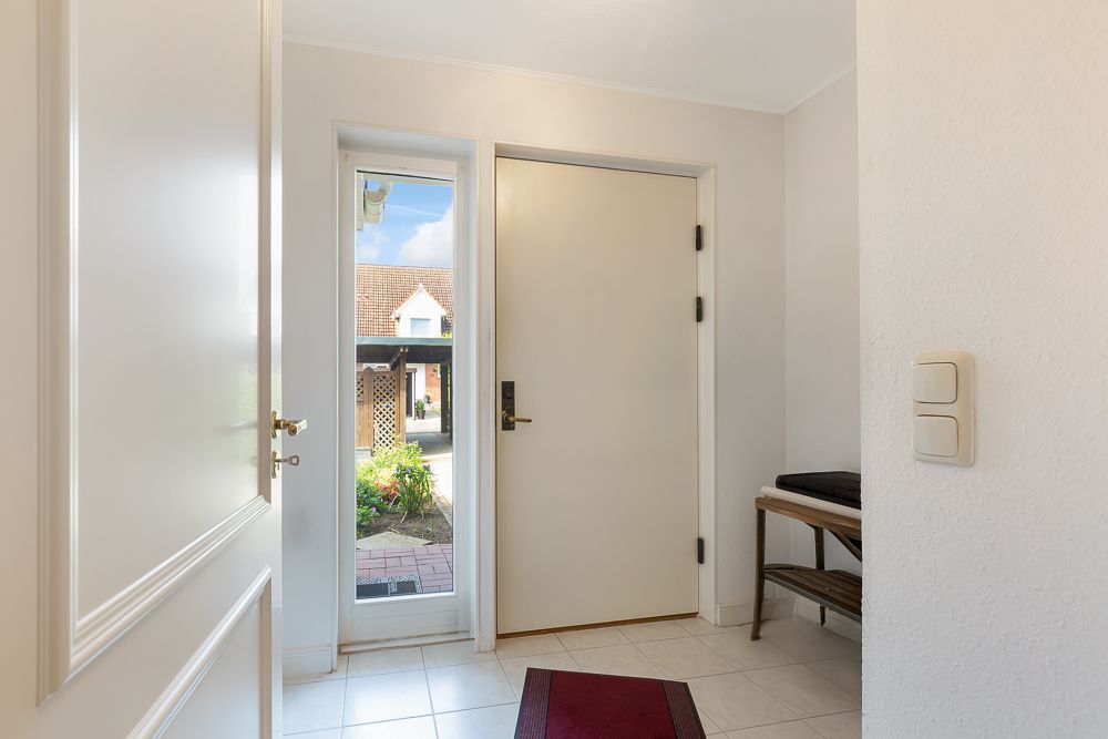 Heller Eingangsbereich mit weißer Tür, Fenster, Fliesenboden und kleinem Regal mit Ablage.