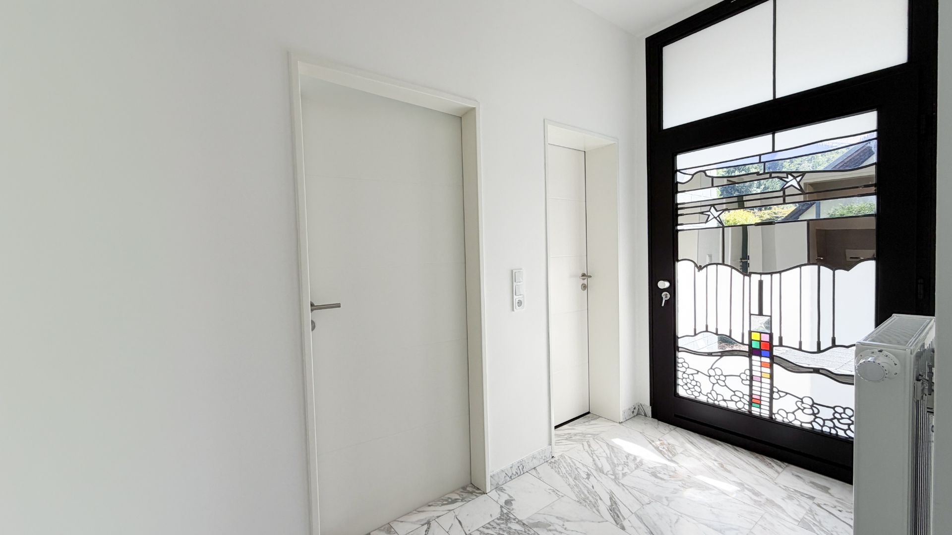 Heller Flur mit Marmorboden, zwei weißen Türen und großer Glastür mit buntem Muster.