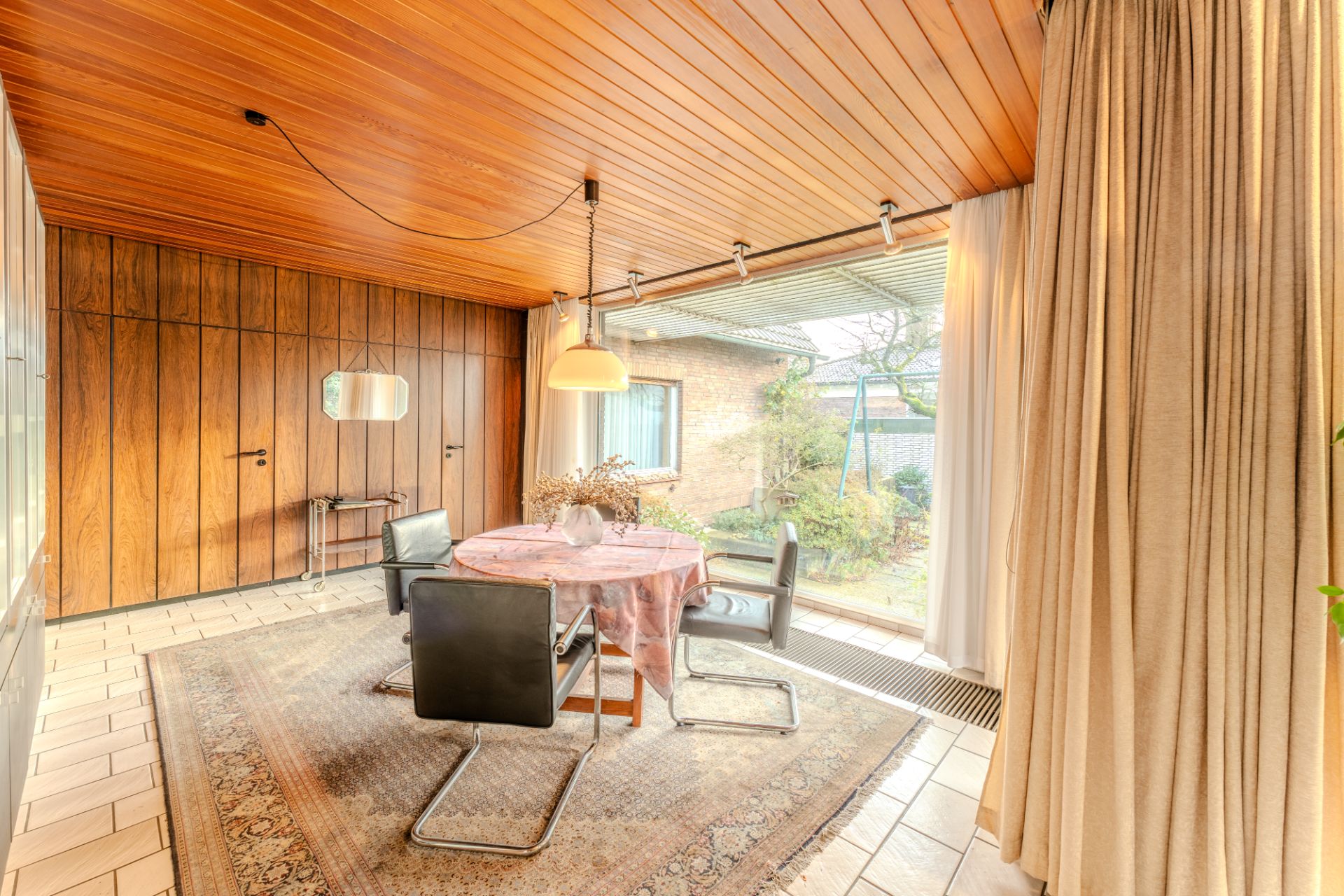 Wohnzimmer mit Holzdecke, rundem Tisch, vier Stühlen und großem Fenster zum Garten.