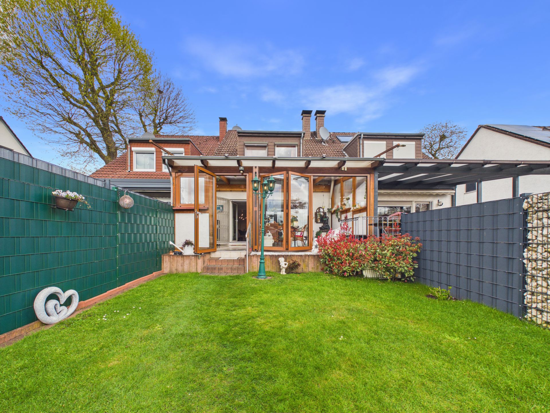 Garten mit grünem Rasen, Terrasse mit Glaswänden und Blumenbeeten hinter einem Haus.