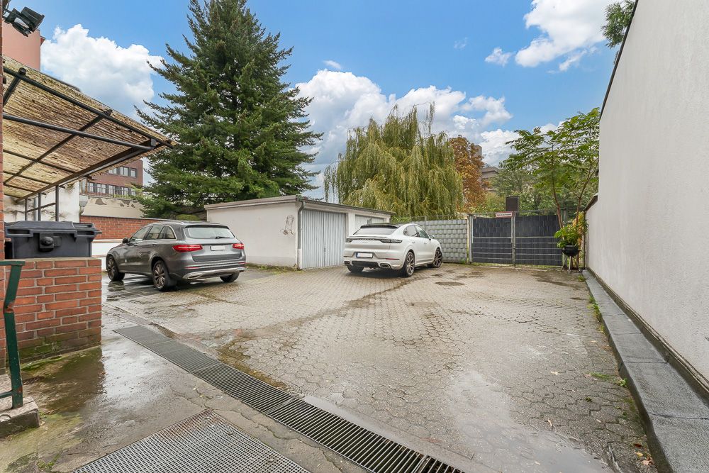 Gepflasterter Hof mit zwei geparkten Autos, Mülltonnen und Bäumen im Hintergrund.