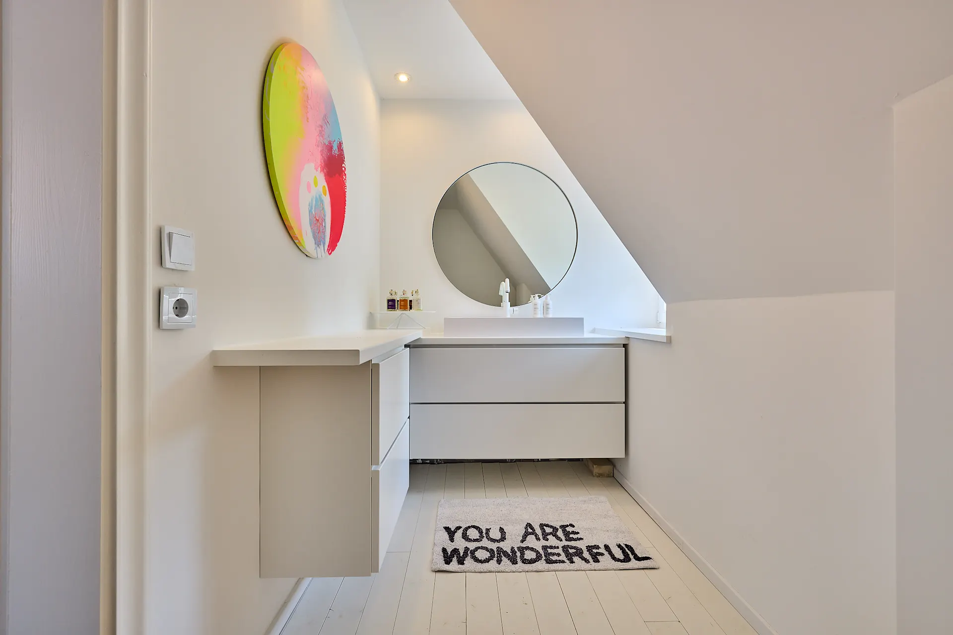 Modernes Badezimmer mit rundem Spiegel, weißem Waschbecken und Teppich mit Aufschrift.