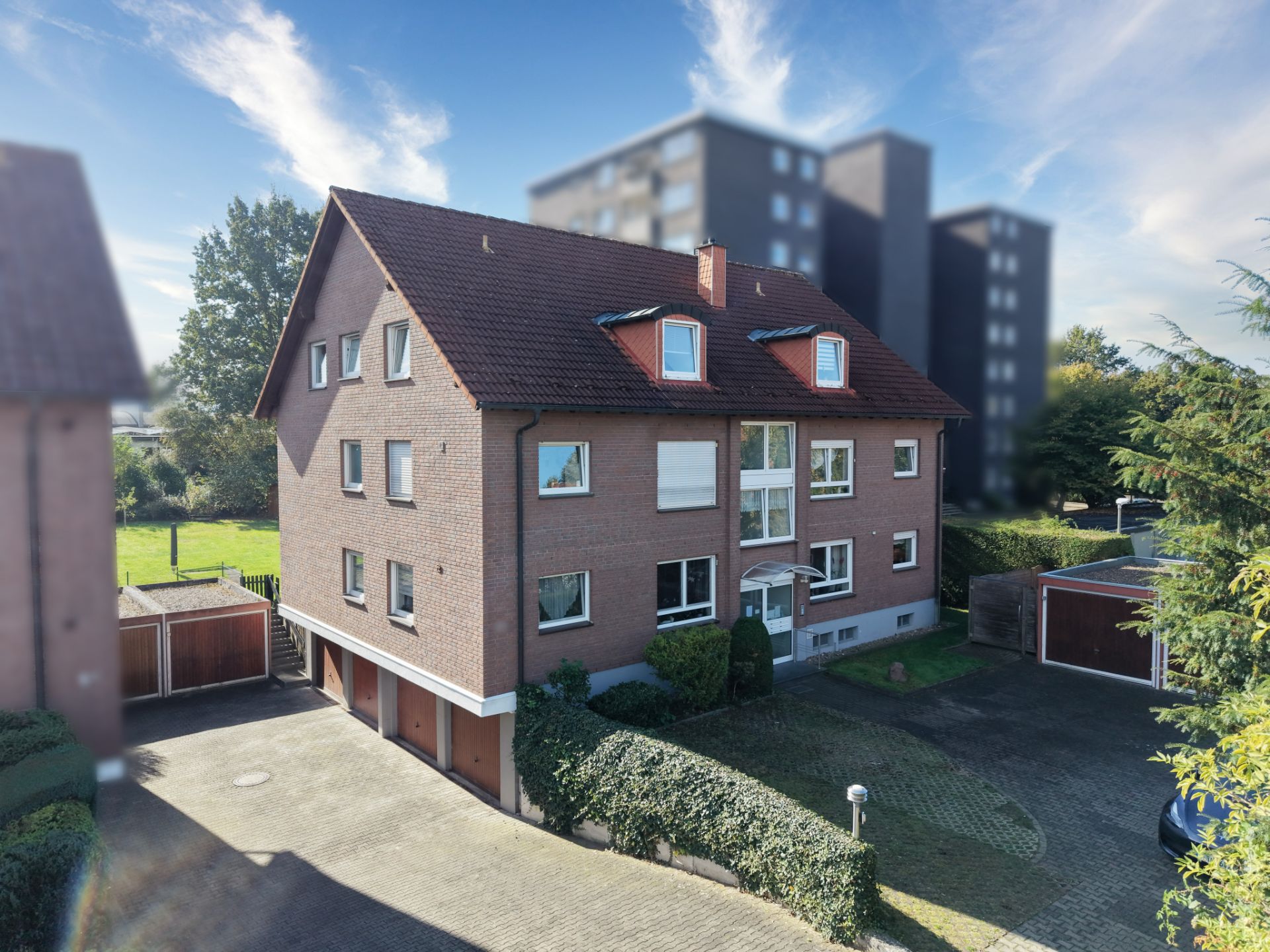 Mehrfamilienhaus aus rotem Backstein mit Satteldach, Garagen und Zufahrt im Vordergrund.