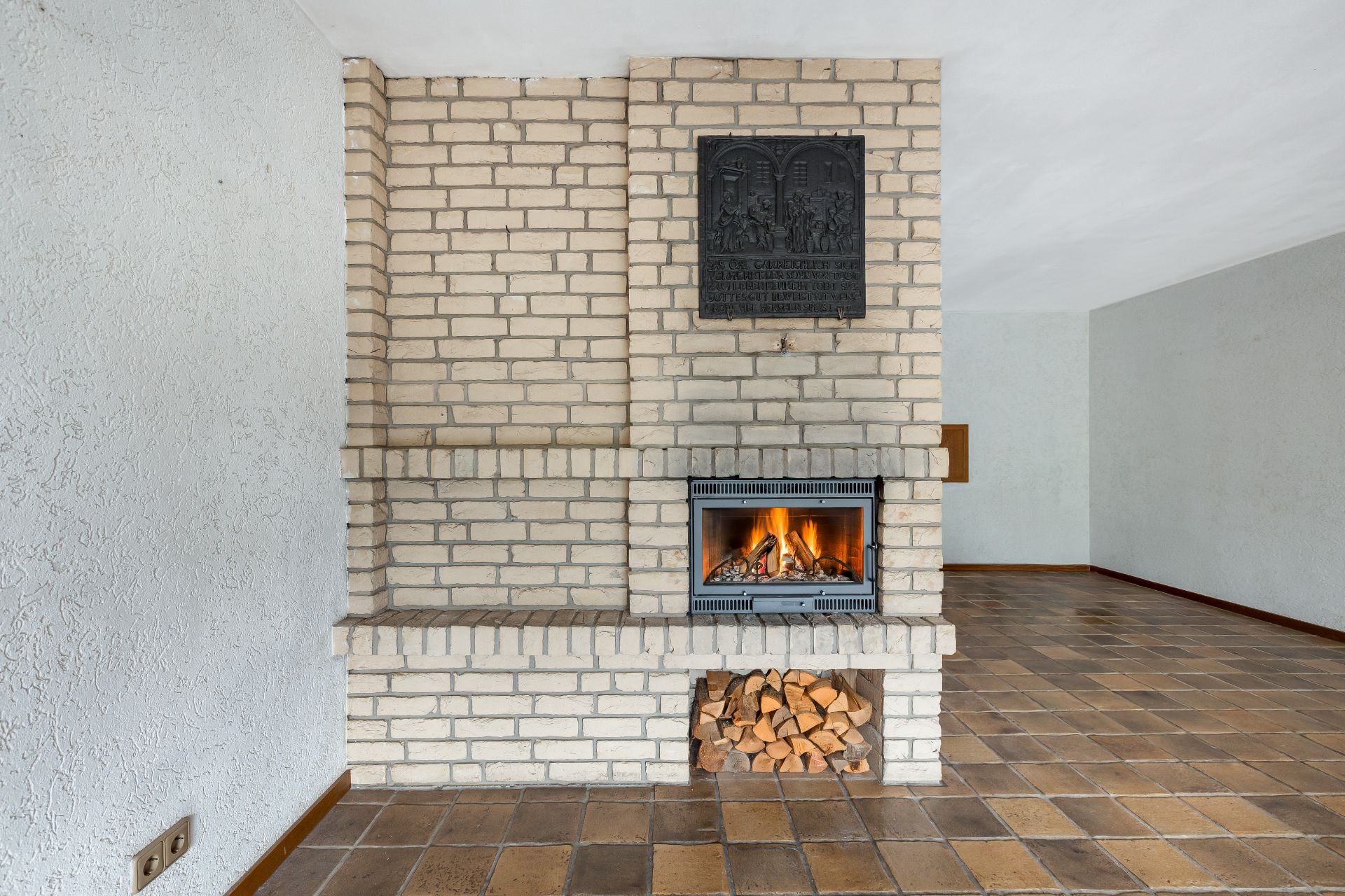 Heller Wohnraum mit gemauertem Kamin, Feuer brennt, Holz darunter gestapelt.