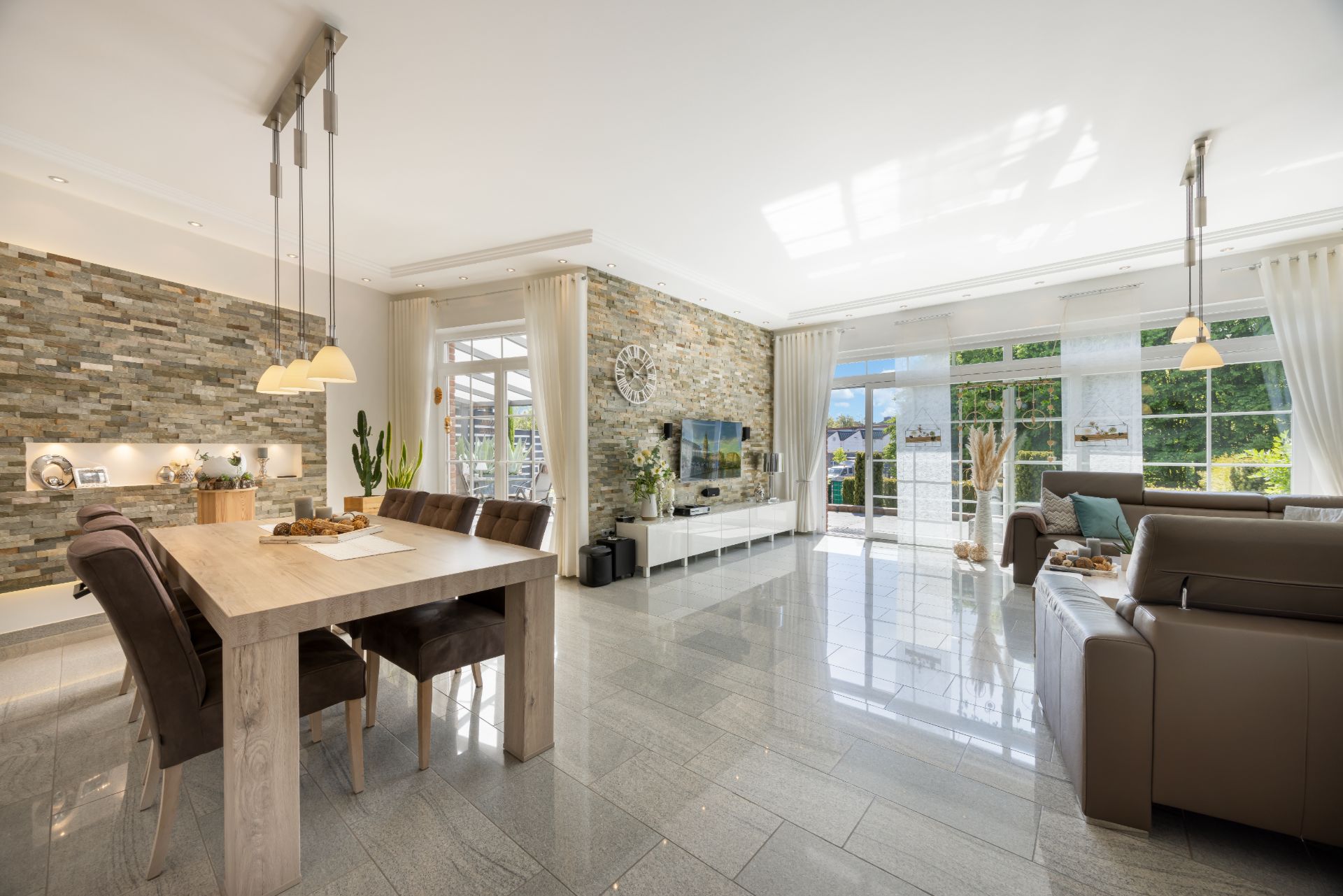 Modernes Wohnzimmer mit Steinwand, Esstisch, Sofas und großen Fenstern mit Vorhängen.