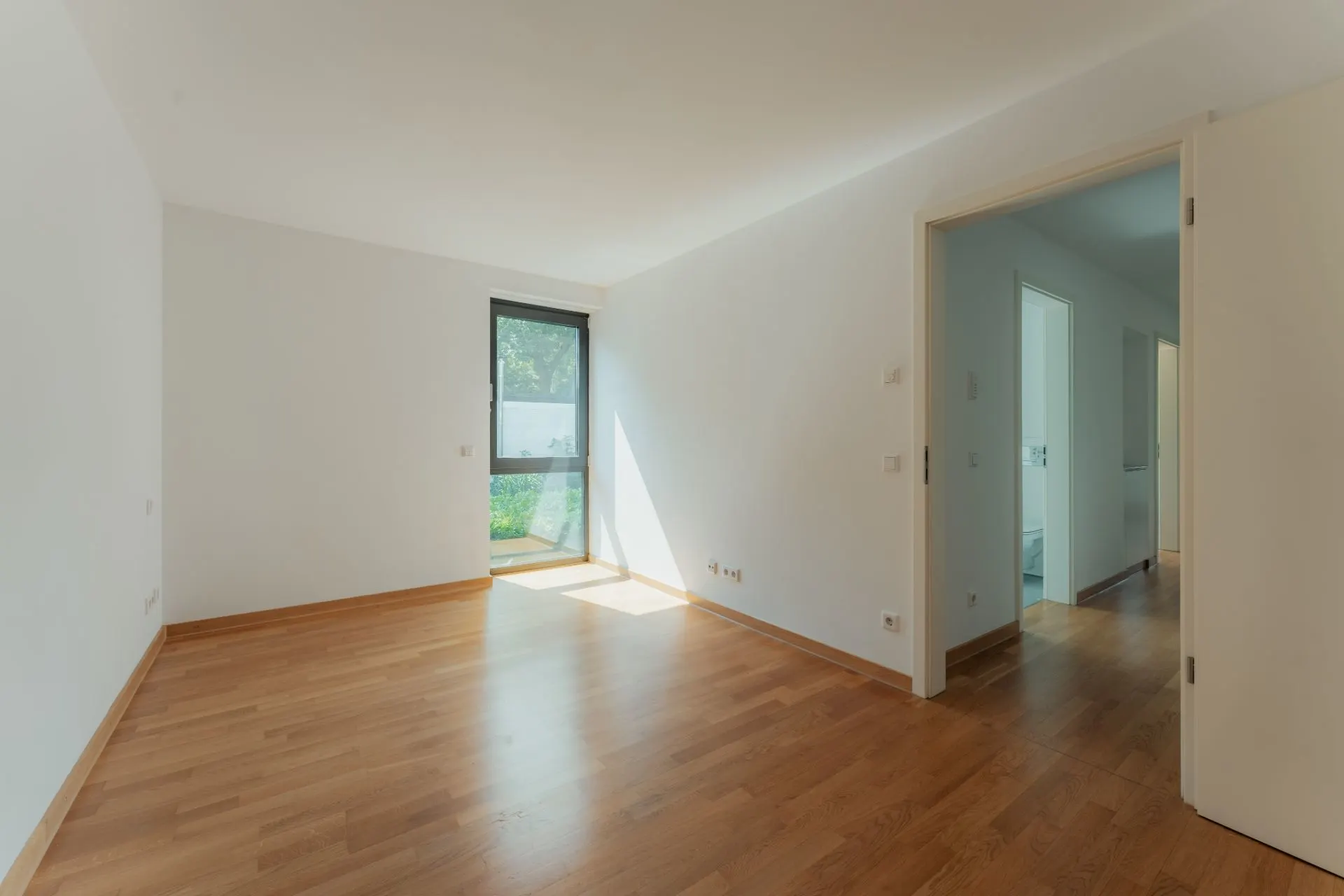 Leerer, heller Raum mit Holzfußboden, Fenster und offener Tür zu einem Flur.