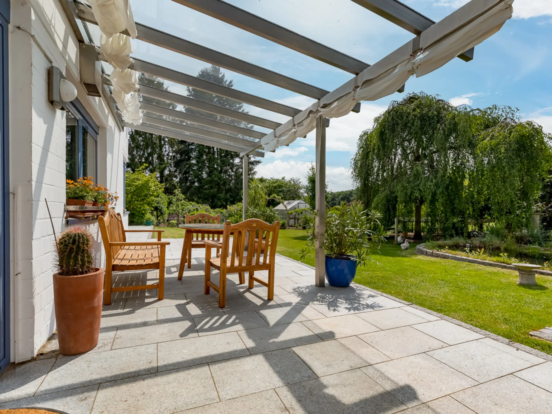 Überdachte Terrasse mit Holzmöbeln, Blumentöpfen und Blick auf einen grünen Garten.