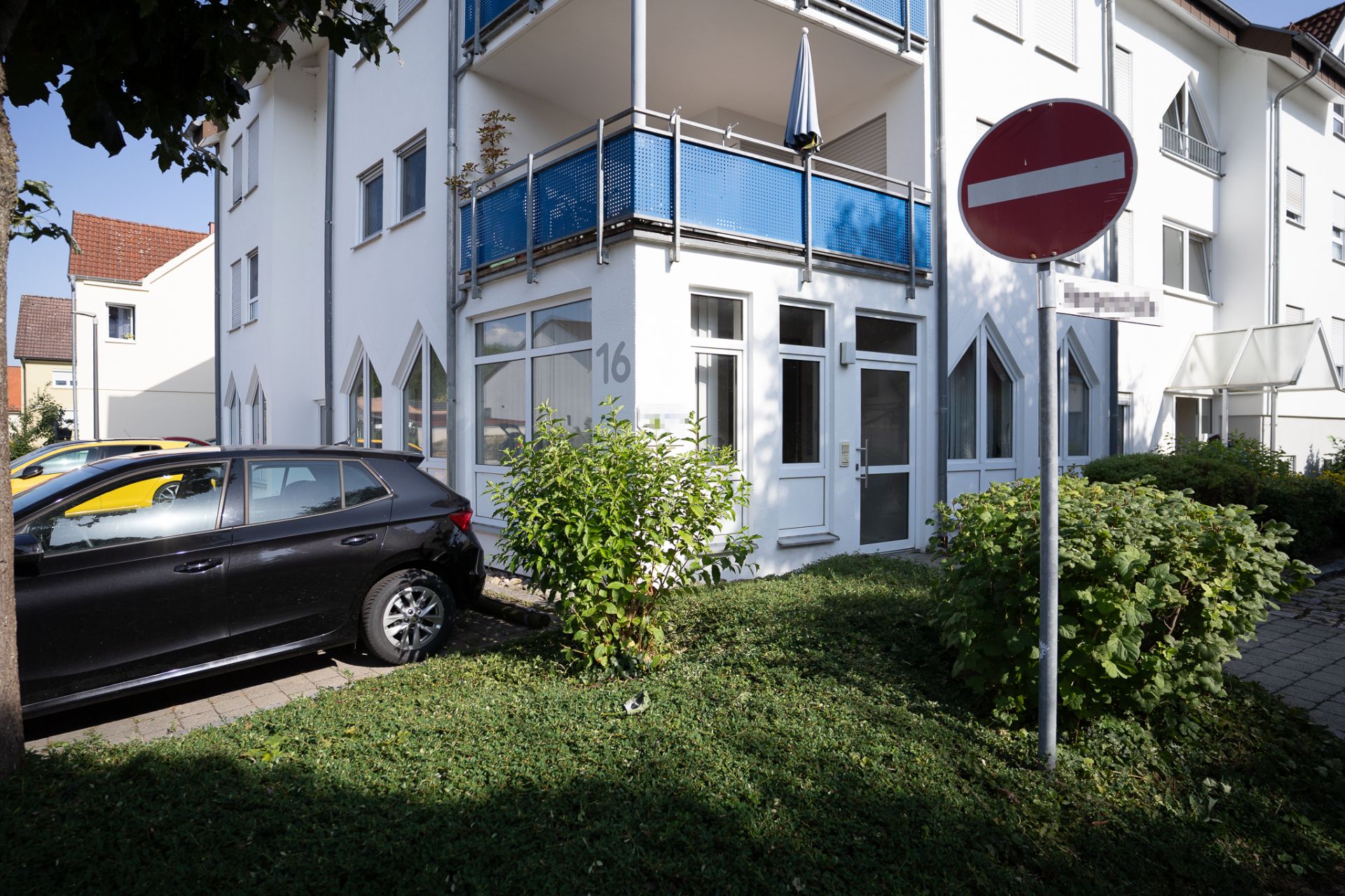 Eingang eines Wohnhauses mit blauen Balkonen, geparktem Auto und Einfahrt-verboten-Schild.