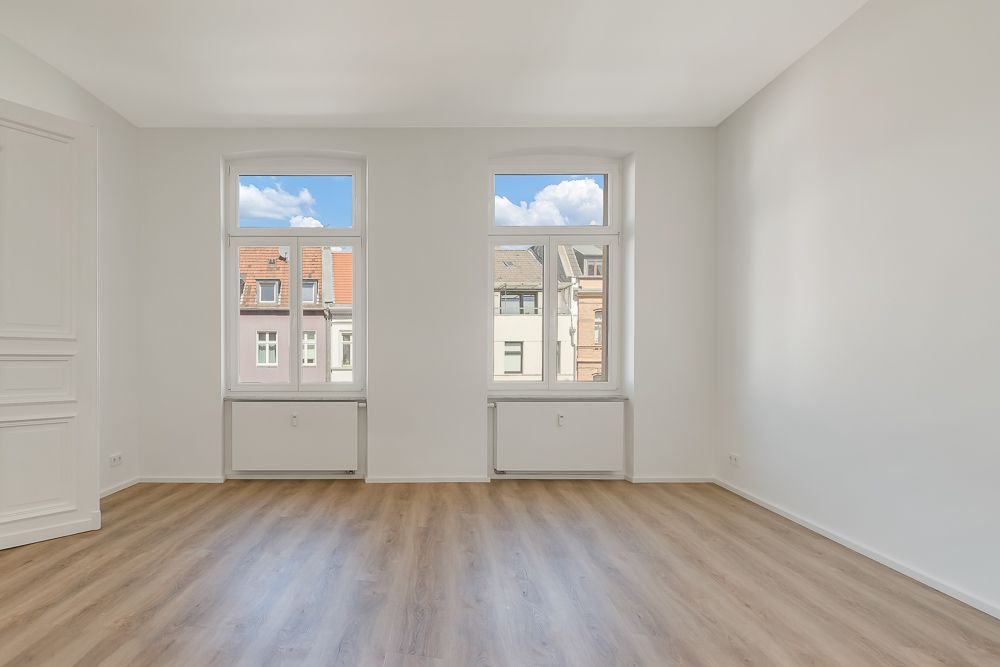 Heller leerer Raum mit zwei großen Fenstern, weißer Wand und Holzboden.