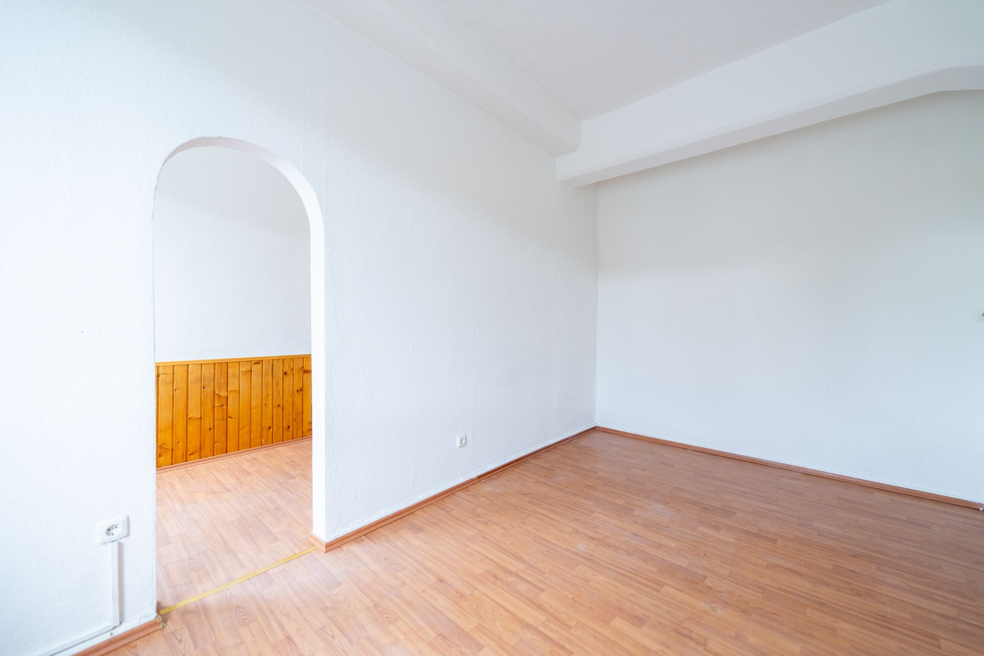 Leerer Raum mit weißer Wand, Holzboden und Rundbogen-Eingang zu einem Nebenraum mit Holzverkleidung.