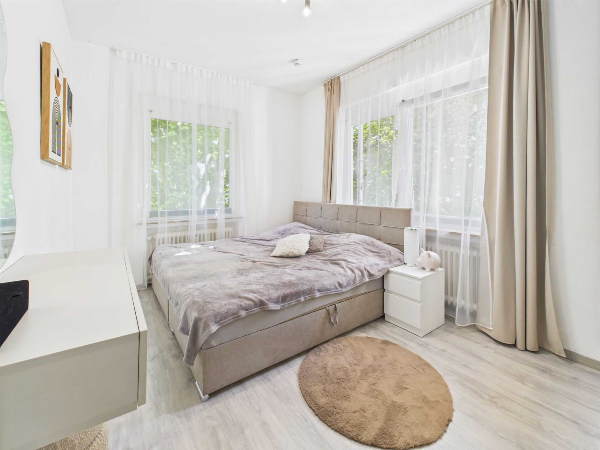 Helles Schlafzimmer mit Doppelbett, Nachttisch, Teppich und großen Fenstern mit Vorhängen.