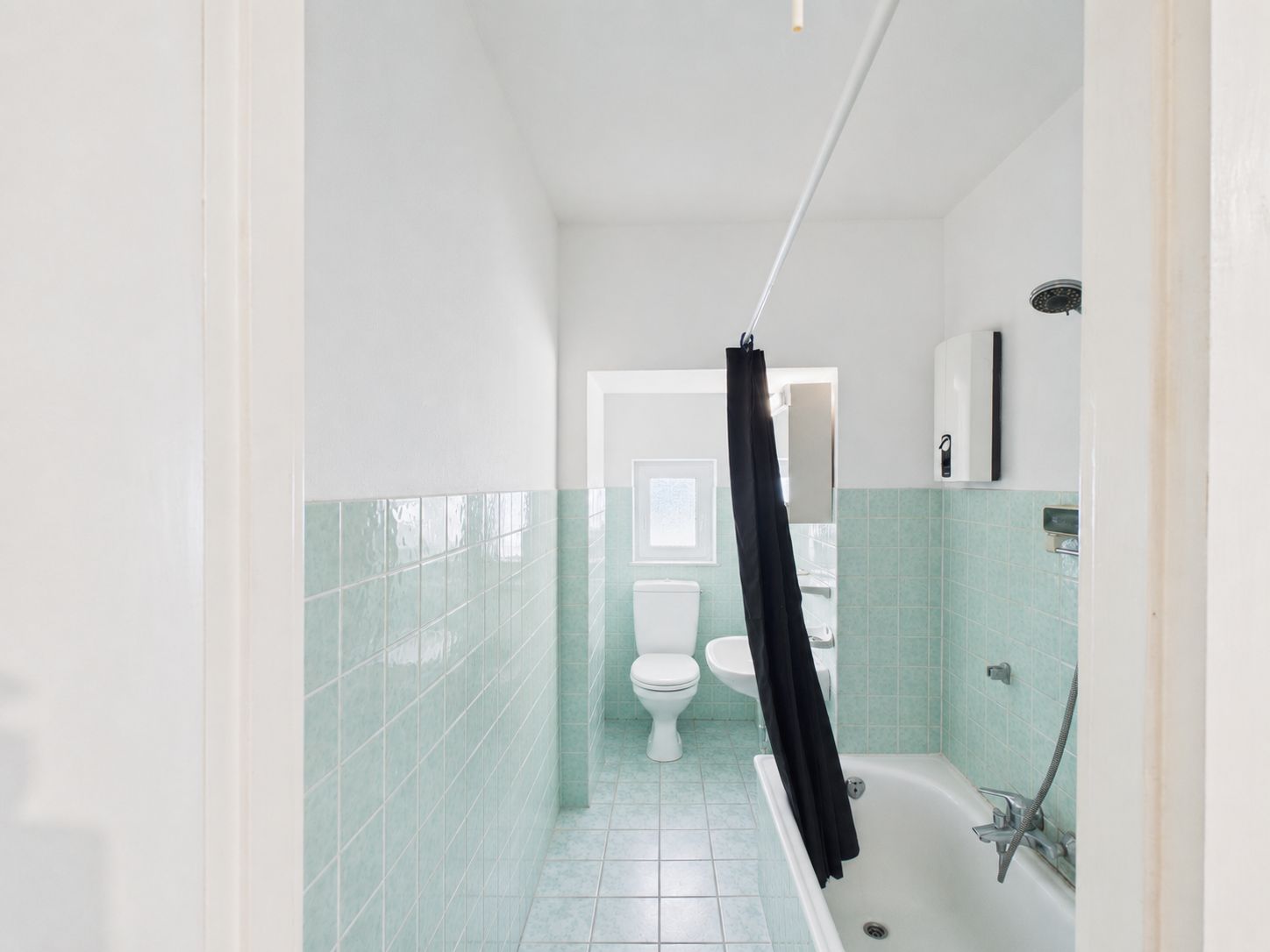 Badezimmer mit grünen Fliesen, Badewanne, Duschvorhang, WC und Fenster.