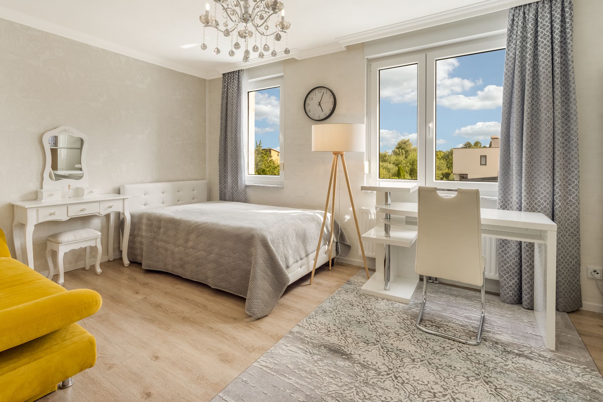 Helles Schlafzimmer mit Bett, Schreibtisch, gelbem Sessel und zwei Fenstern mit Vorhängen.