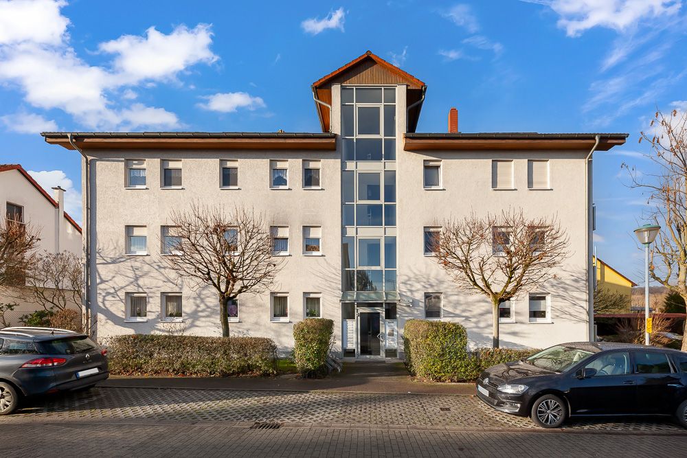 Dreistöckiges Wohnhaus mit zentralem gläsernen Treppenhaus, geparkte Autos und Bäume davor.