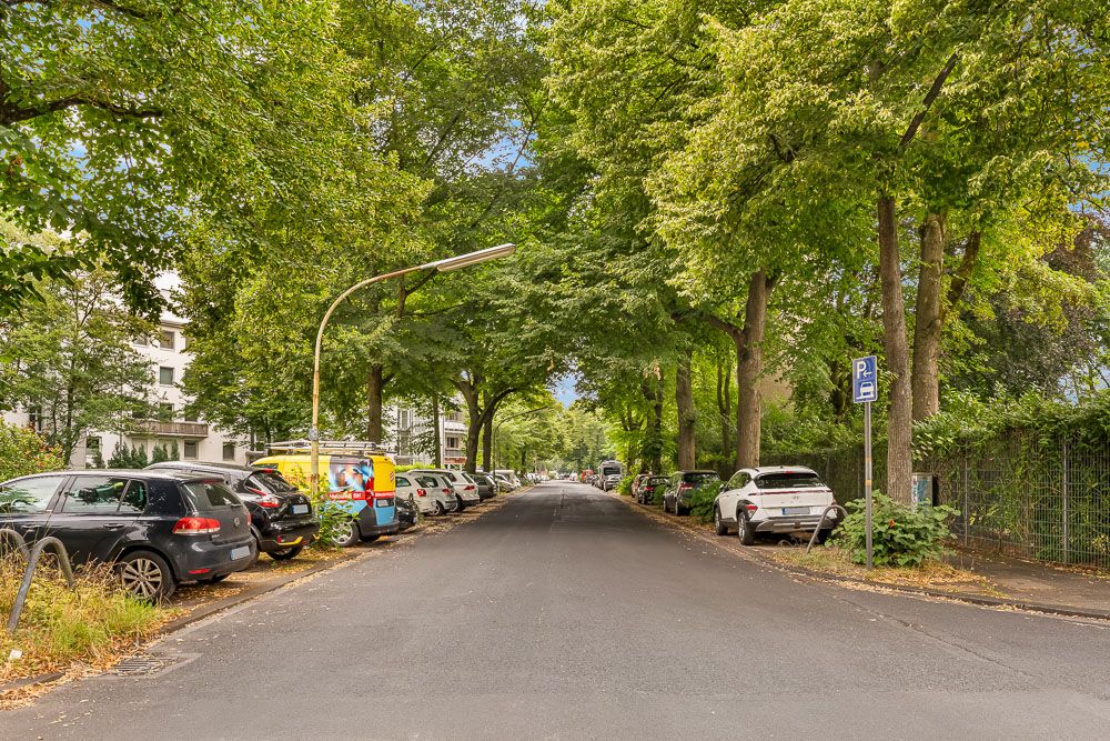 Baumbestandene Straße mit parkenden Autos an beiden Seiten, Wohnhäuser links im Bild.