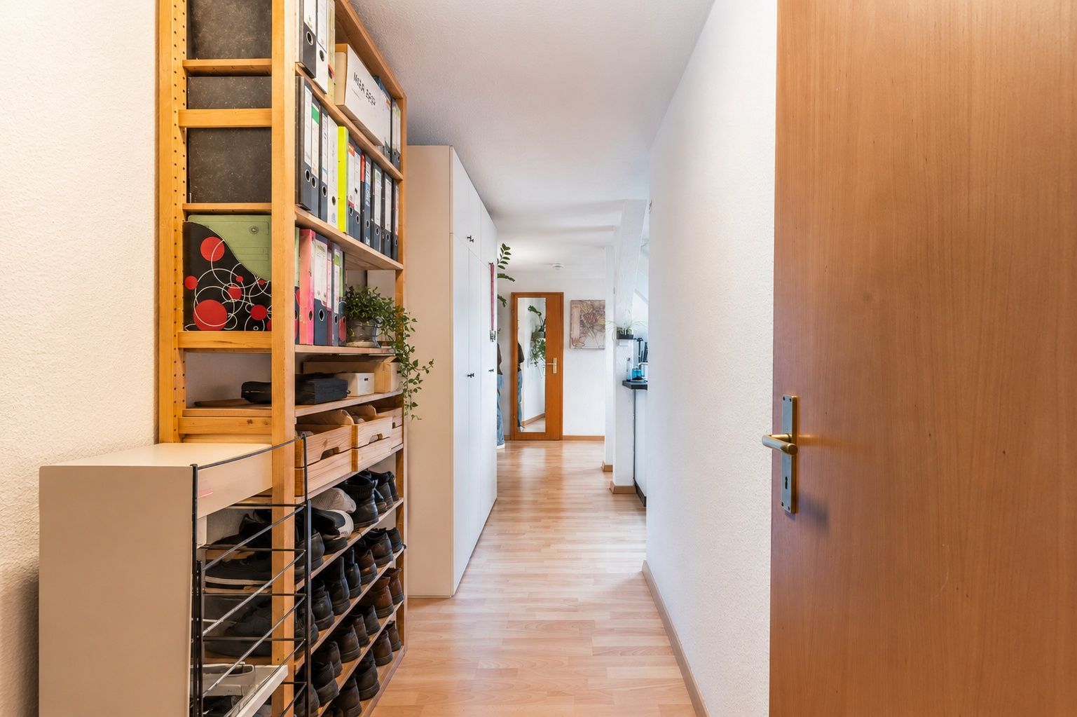 Heller Flur mit Holzregal voller Schuhe, Aktenordner und Pflanzen, daneben eine offene Holztür.