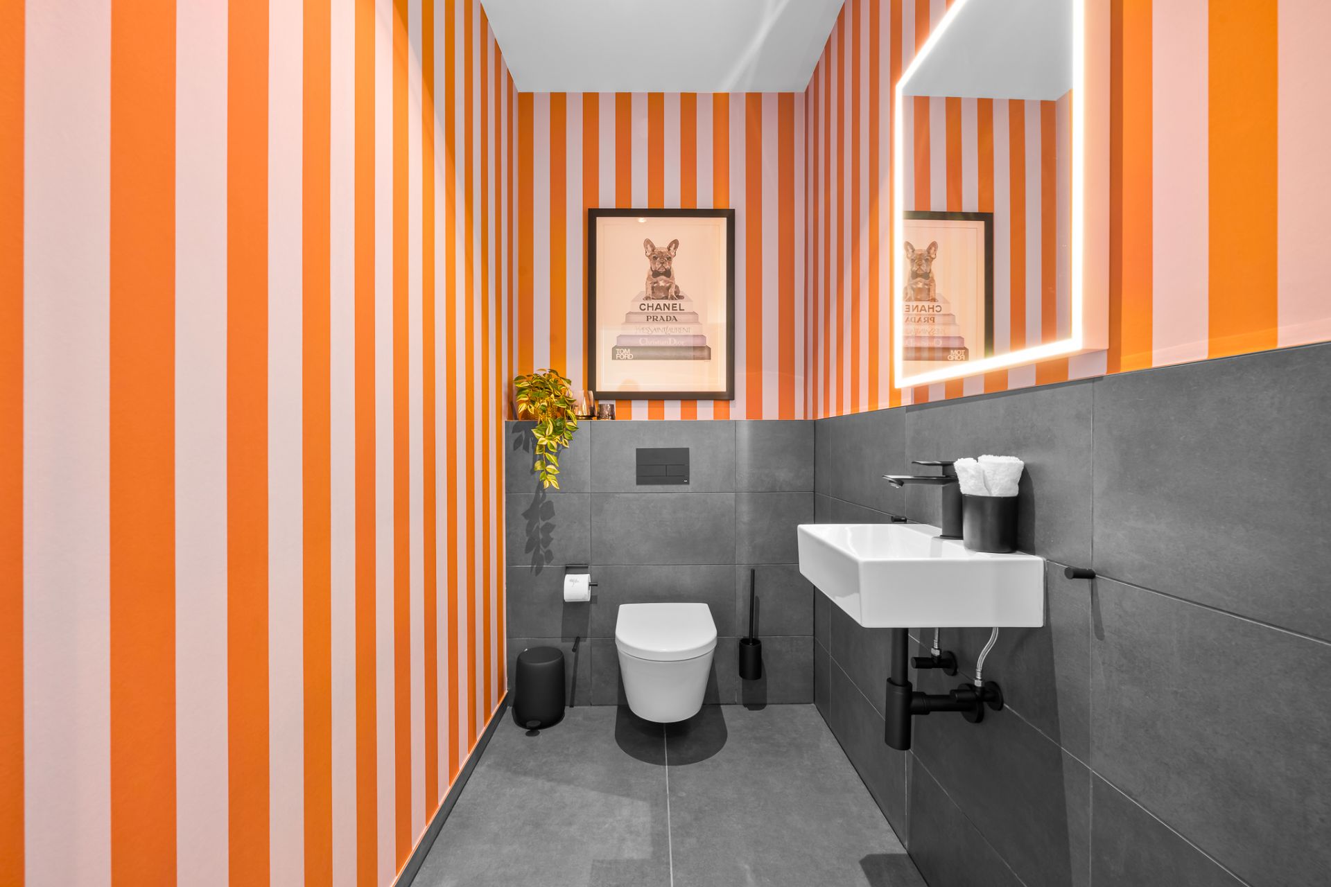 Modernes WC mit weißem Waschbecken, beleuchtetem Spiegel und orange-weiß gestreifter Tapete.