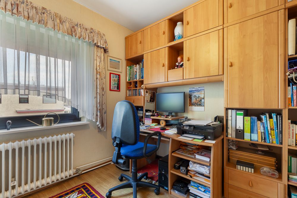 Kleines Arbeitszimmer mit Schreibtisch, Computer, Regal, Bücher und Fenster mit Vorhang.