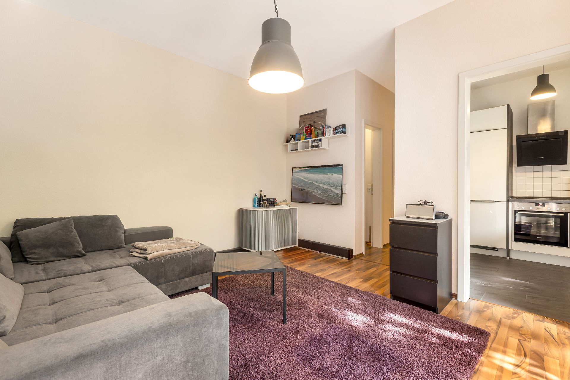 Wohnzimmer mit grauem Ecksofa, Couchtisch, violettem Teppich und Blick in die Küche.