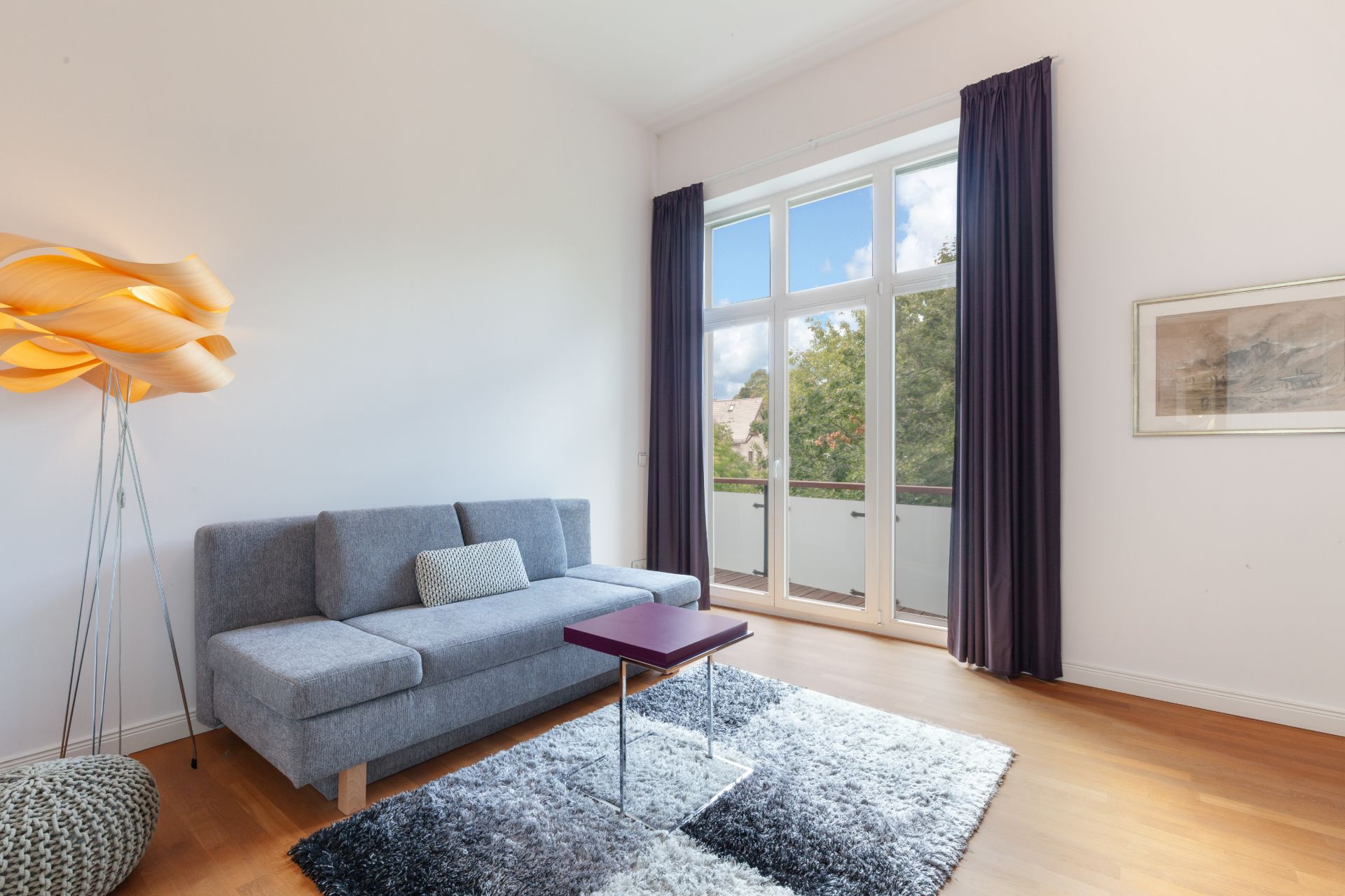 Helles Wohnzimmer mit grauem Sofa, Stehlampe, Teppich und Zugang zum Balkon.