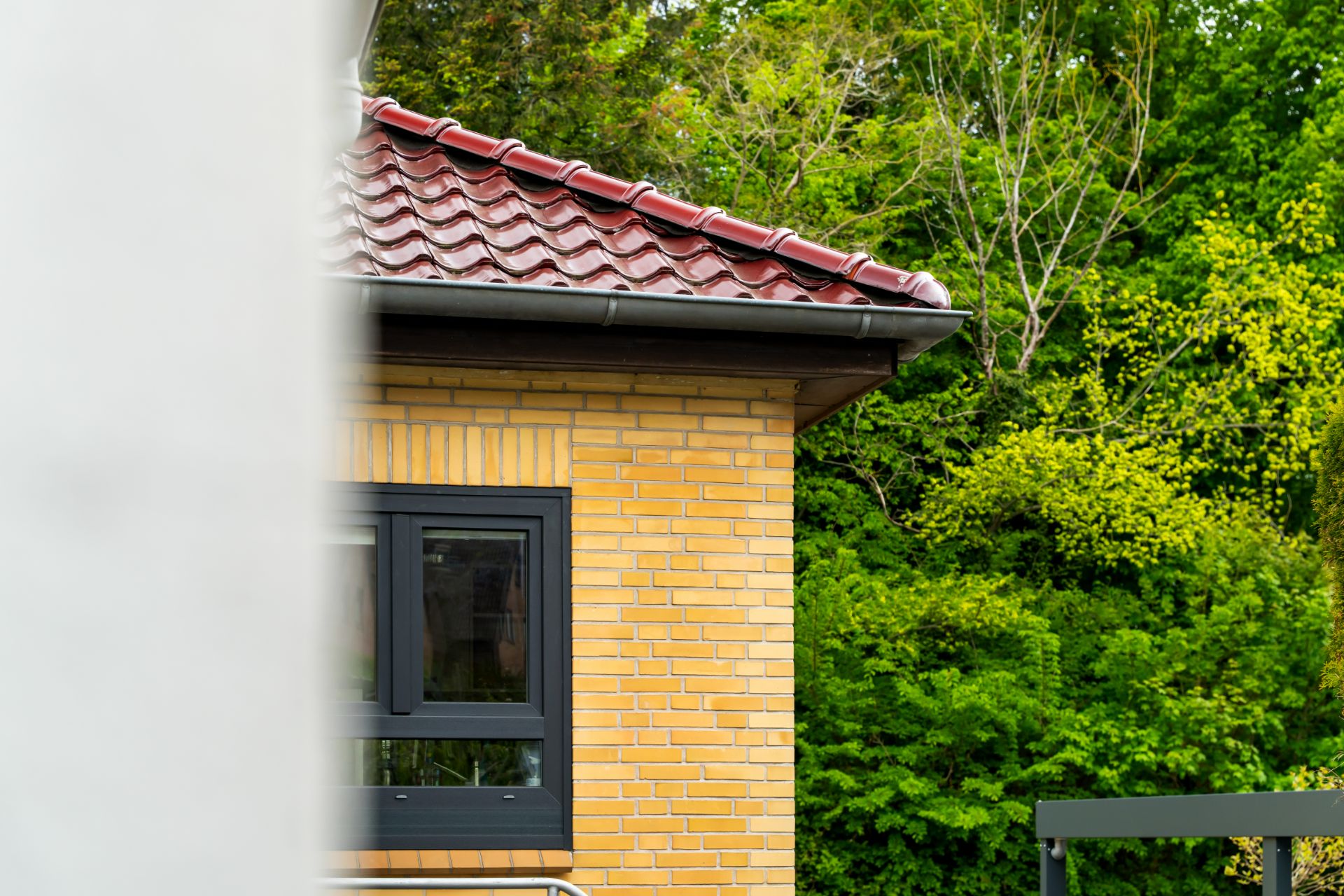 Teil einer gelben Backsteinfassade mit Fenster und rotem Dach, daneben grüne Bäume.
