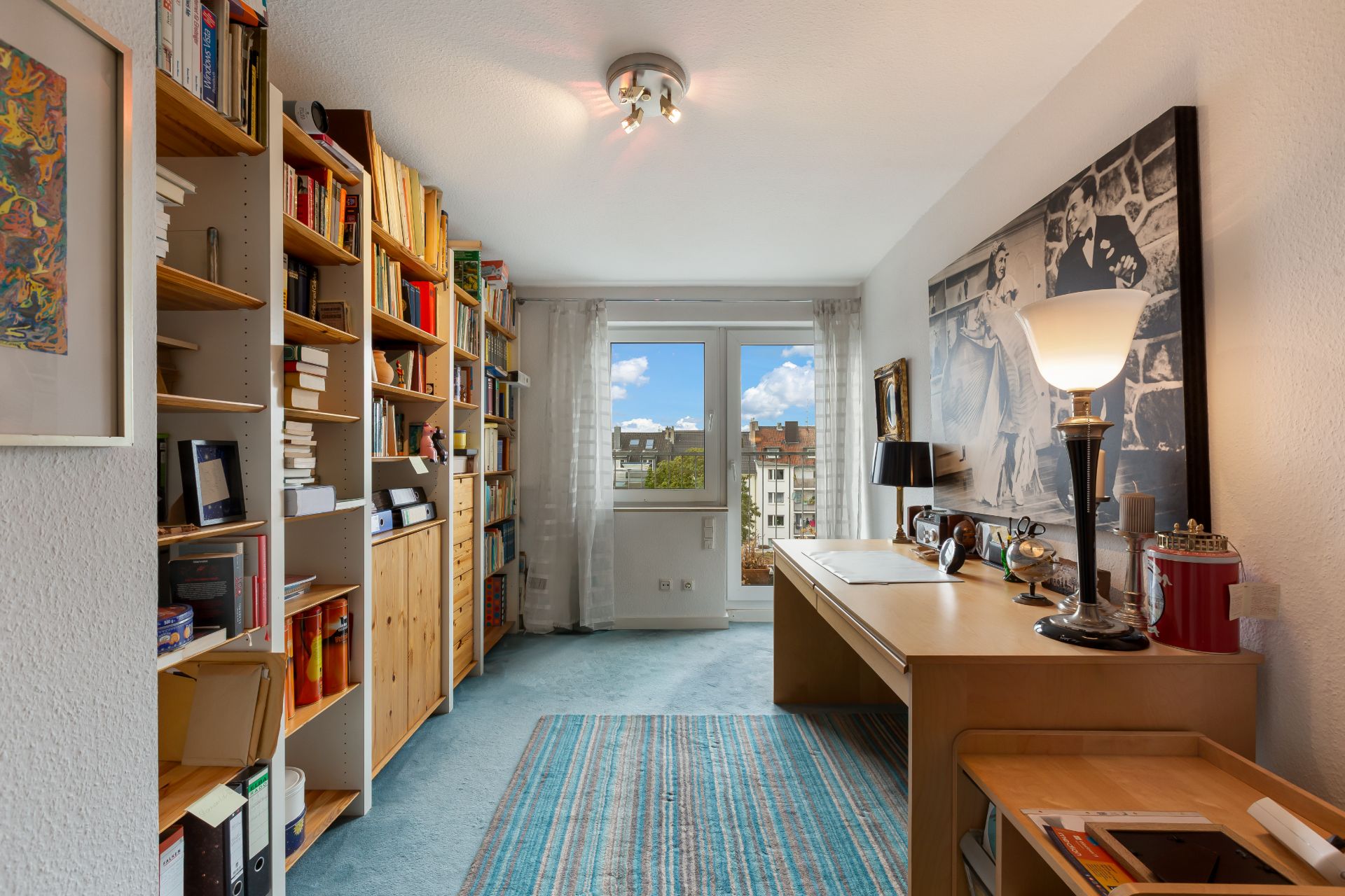 Helles Arbeitszimmer mit Bücherregalen, Schreibtisch, Teppich und großem Fenster mit Blick nach draußen.