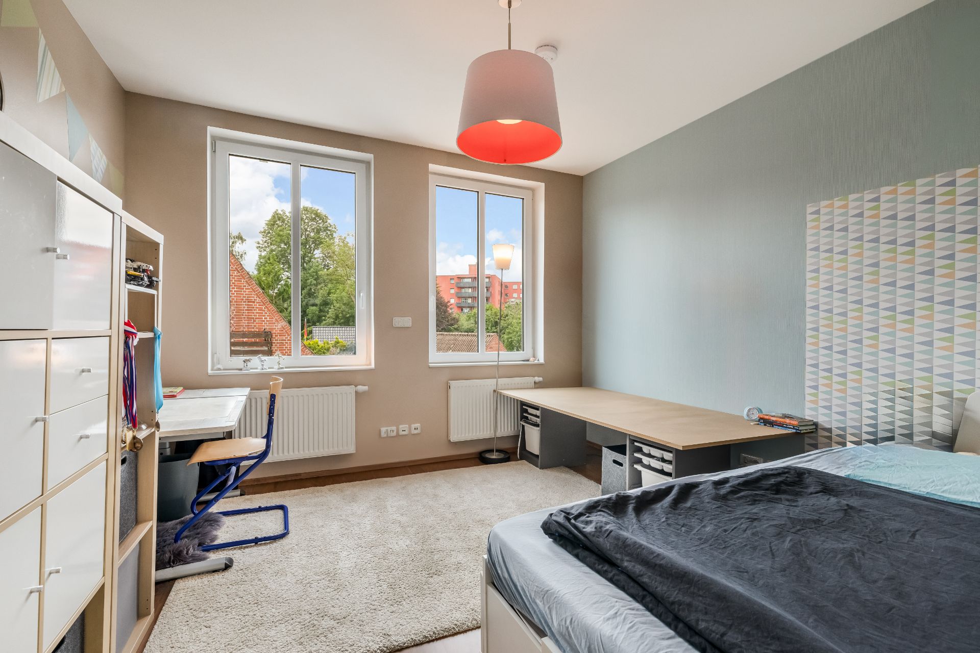 Helles Schlafzimmer mit Bett, Schreibtisch, Regal und zwei Fenstern mit Blick ins Grüne.