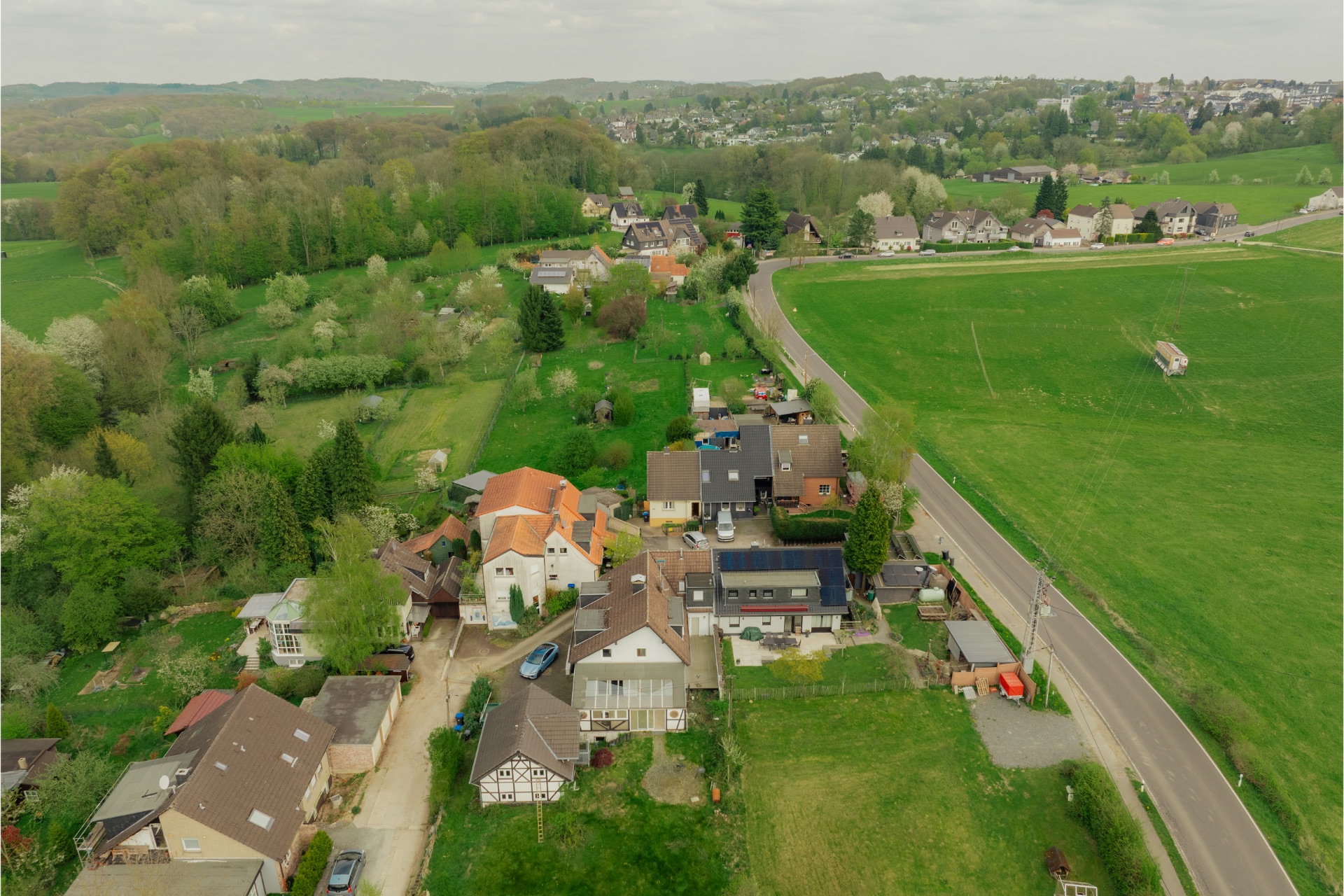 Luftaufnahme von Wohnhäusern mit Gärten am Rand einer Landstraße, umgeben von Feldern und Bäumen.