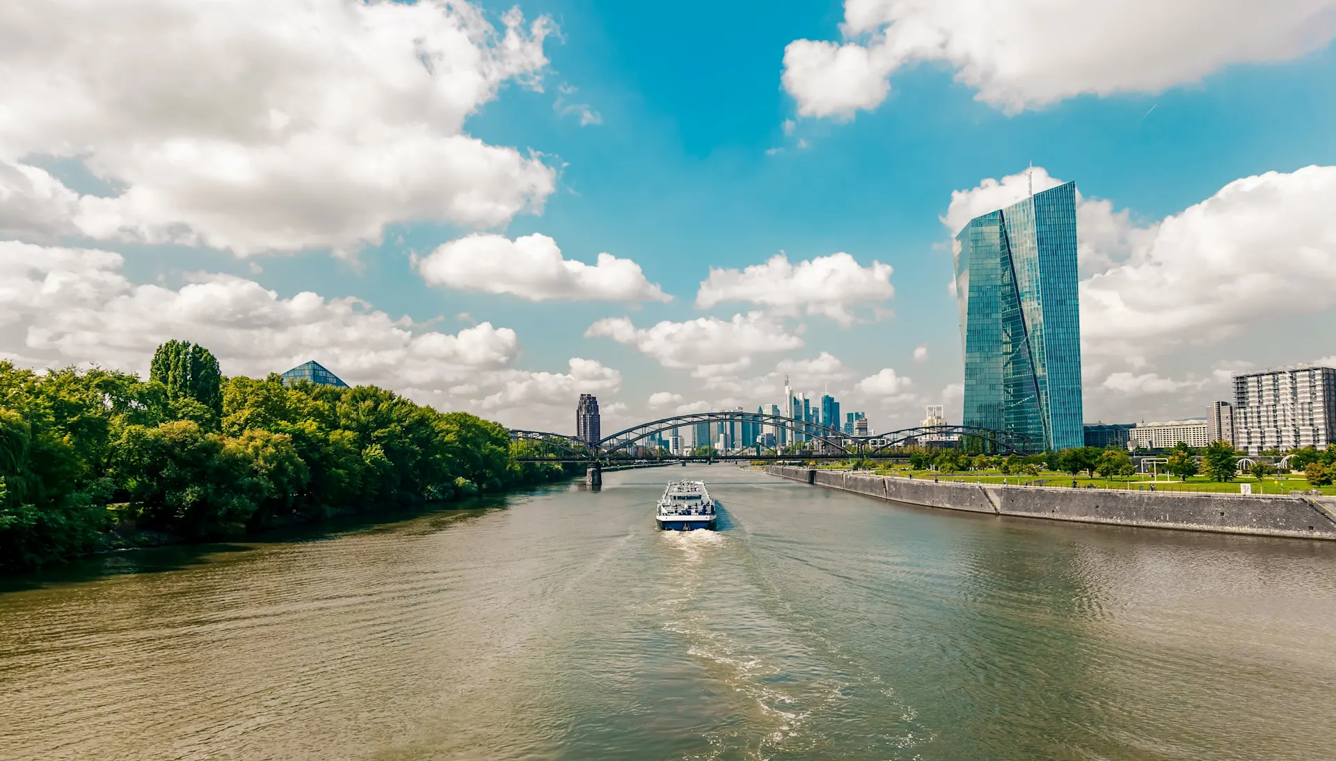 Fluss mit Boot, Brücke im Hintergrund, moderne Hochhäuser und grüne Ufer bei Sonnenschein.