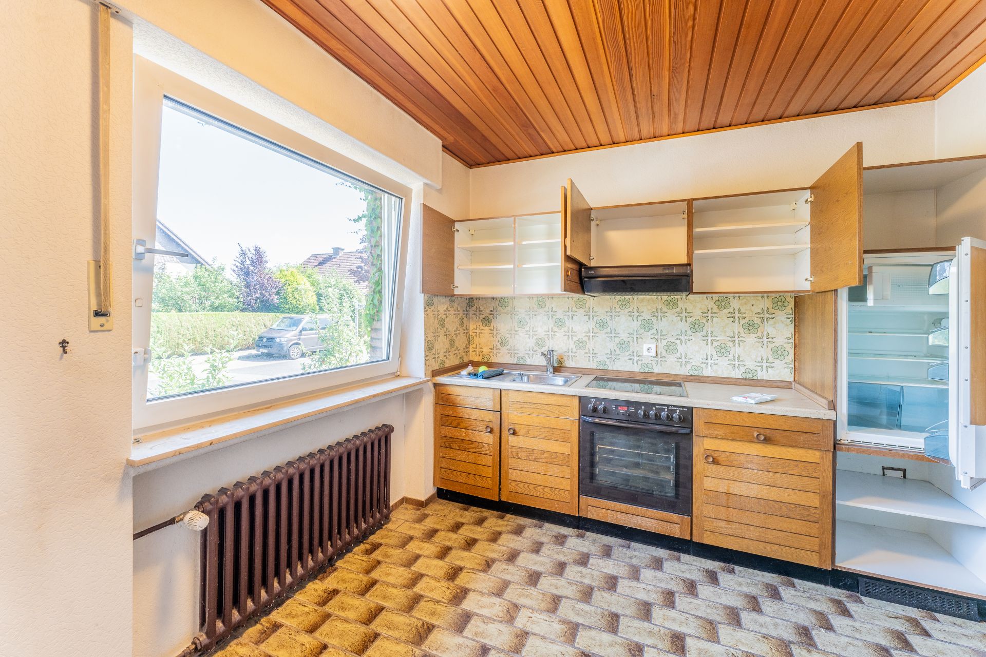 Helle Küche mit Holzdecke, offenem Kühlschrank und Schränken, großem Fenster und gemusterten Fliesen.
