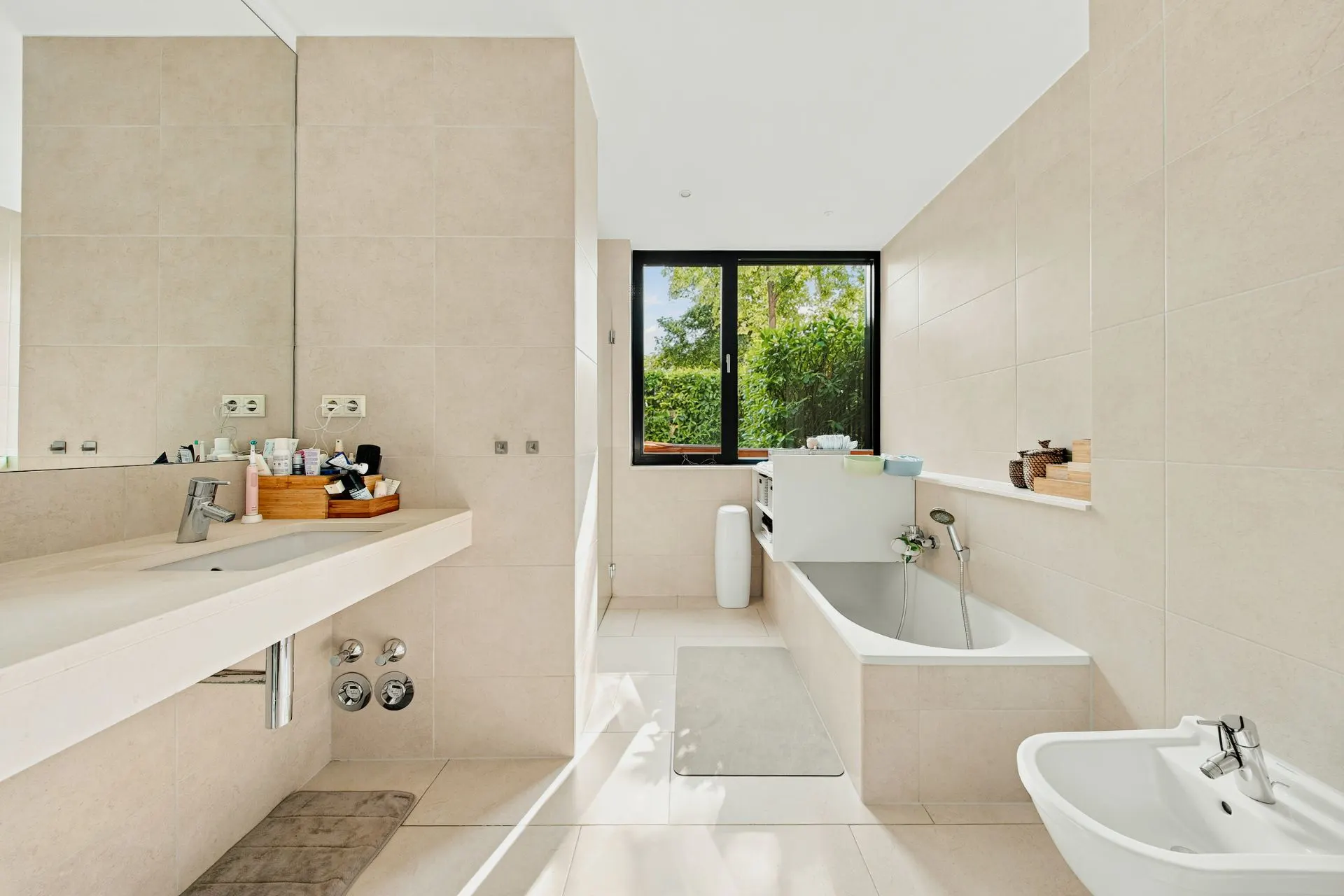 Helles Badezimmer mit Badewanne, Waschbecken, Bidet, großem Spiegel und Fenster mit Blick ins Grüne.
