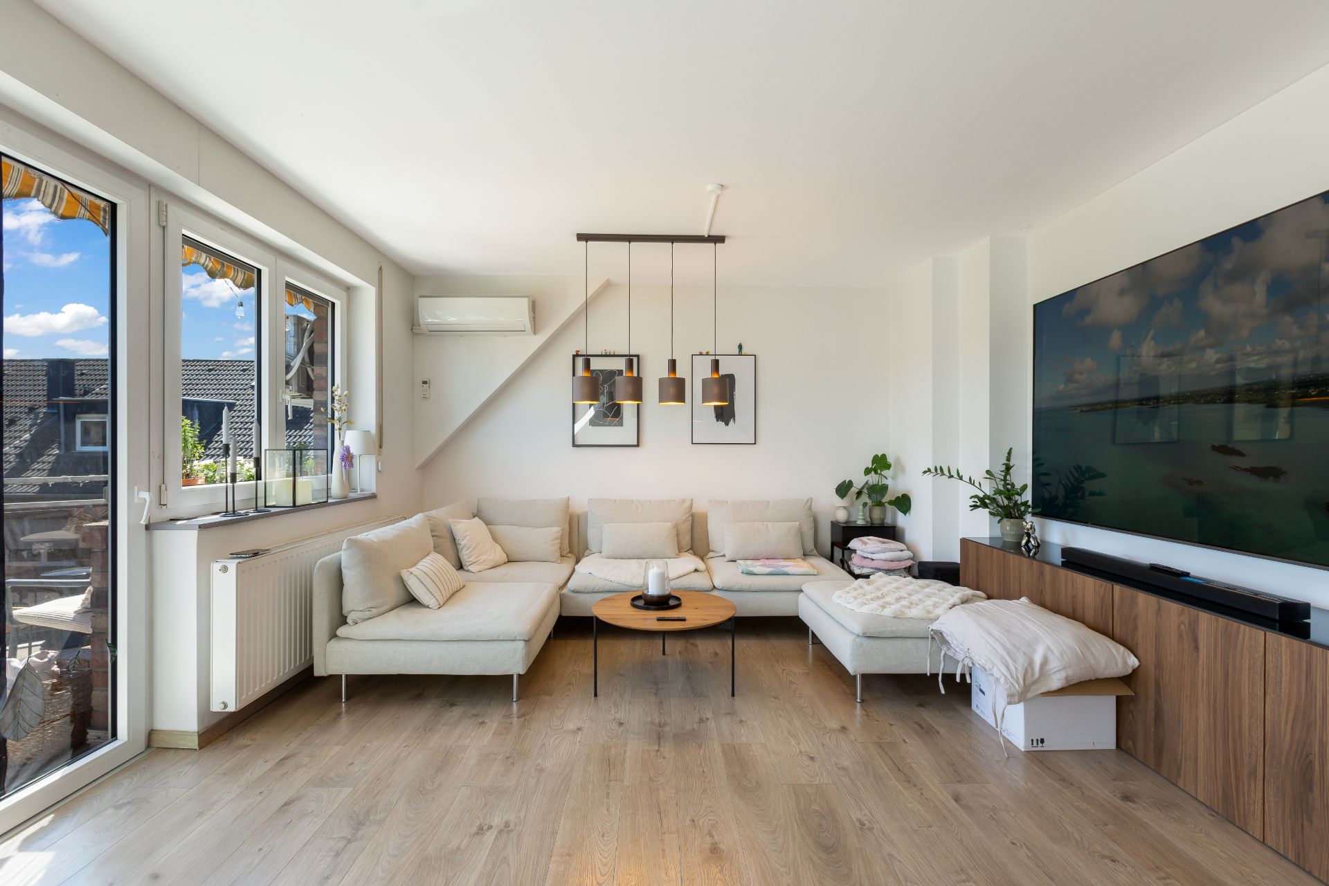 Helles Wohnzimmer mit großem Ecksofa, Couchtisch und großem Fernseher an der Wand.