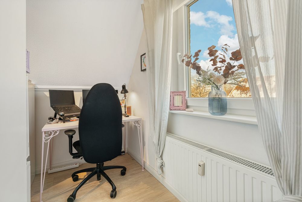Heller Arbeitsbereich mit Schreibtisch, Laptop, Stuhl und Fenster mit Vorhängen und Blumenvase.