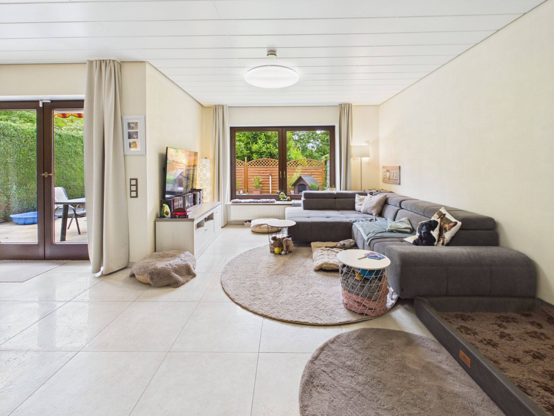Helles Wohnzimmer mit großer Eckcouch, Couchtisch, Teppichen und bodentiefen Fenstern zum Garten.