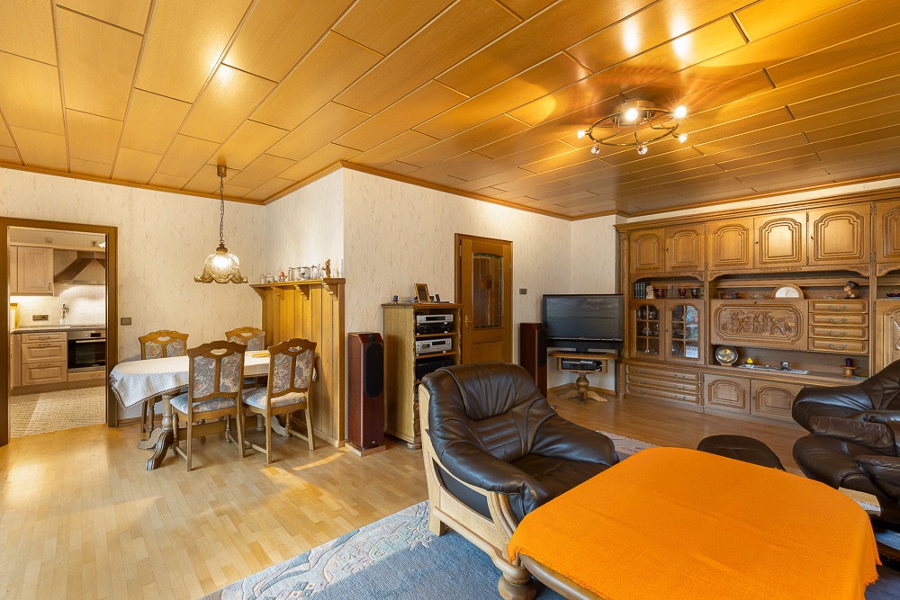 Wohnzimmer mit Holzmöbeln, Essbereich, Ledersofas und Regalwand, angrenzende Küche sichtbar.
