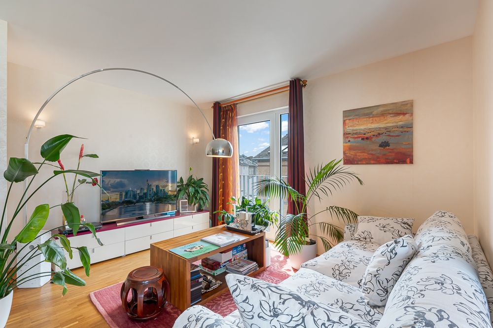 Helles Wohnzimmer mit Muster-Sofa, Couchtisch, großen Pflanzen, Fernseher und Gemälde an der Wand.