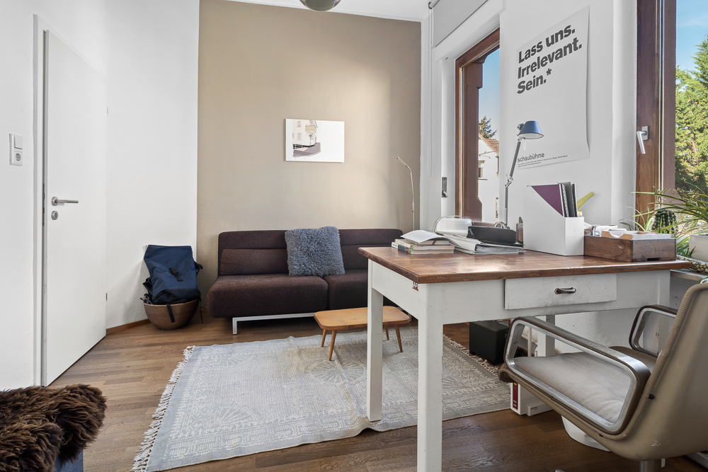 Helles Arbeitszimmer mit Schreibtisch, braunem Sofa, Teppich und großem Fenster.