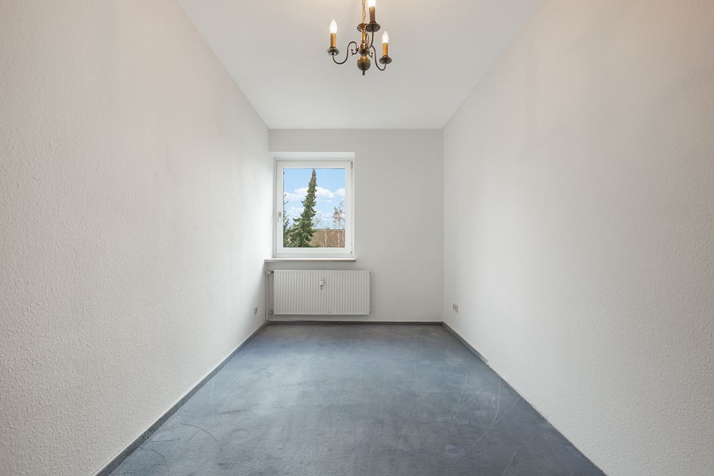Leerer, schmaler Raum mit grauem Teppichboden, weißem Fenster und kleiner Deckenlampe.