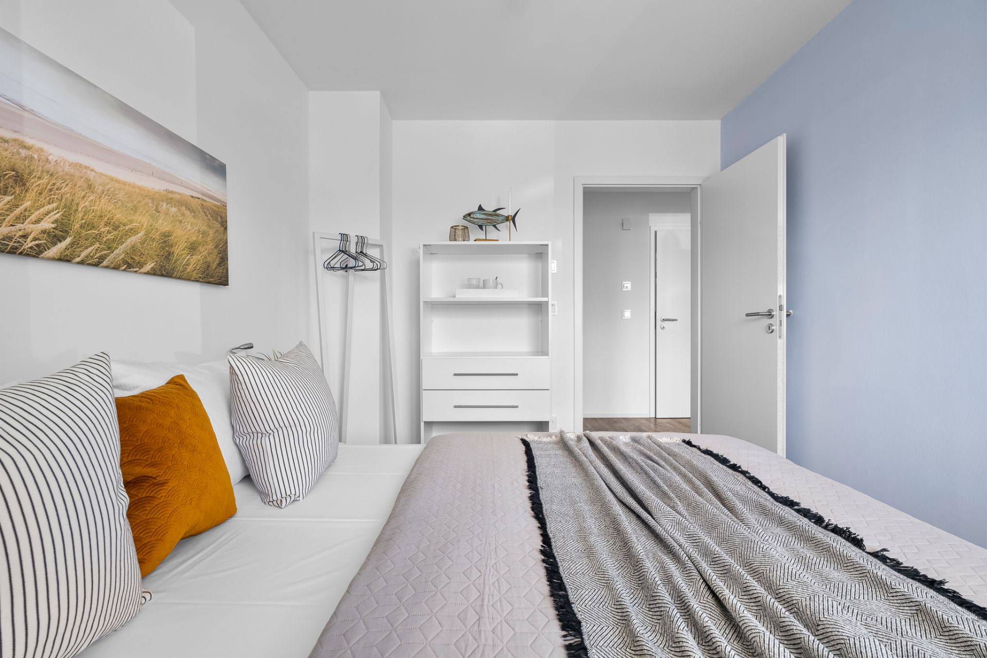 Helles Schlafzimmer mit Bett, Zierkissen, offenem Regal und geöffneter Tür.