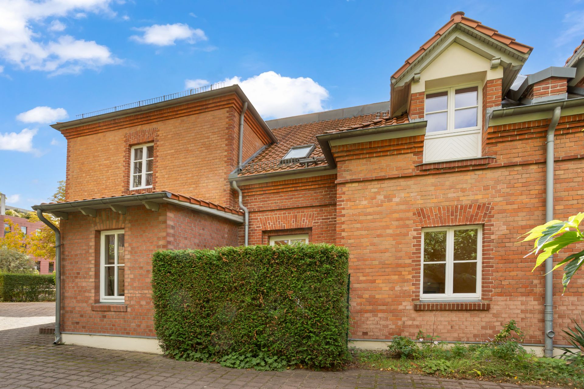 Backsteinwohnhaus mit mehreren Fenstern, Hecken und gepflastertem Hof im Vordergrund.