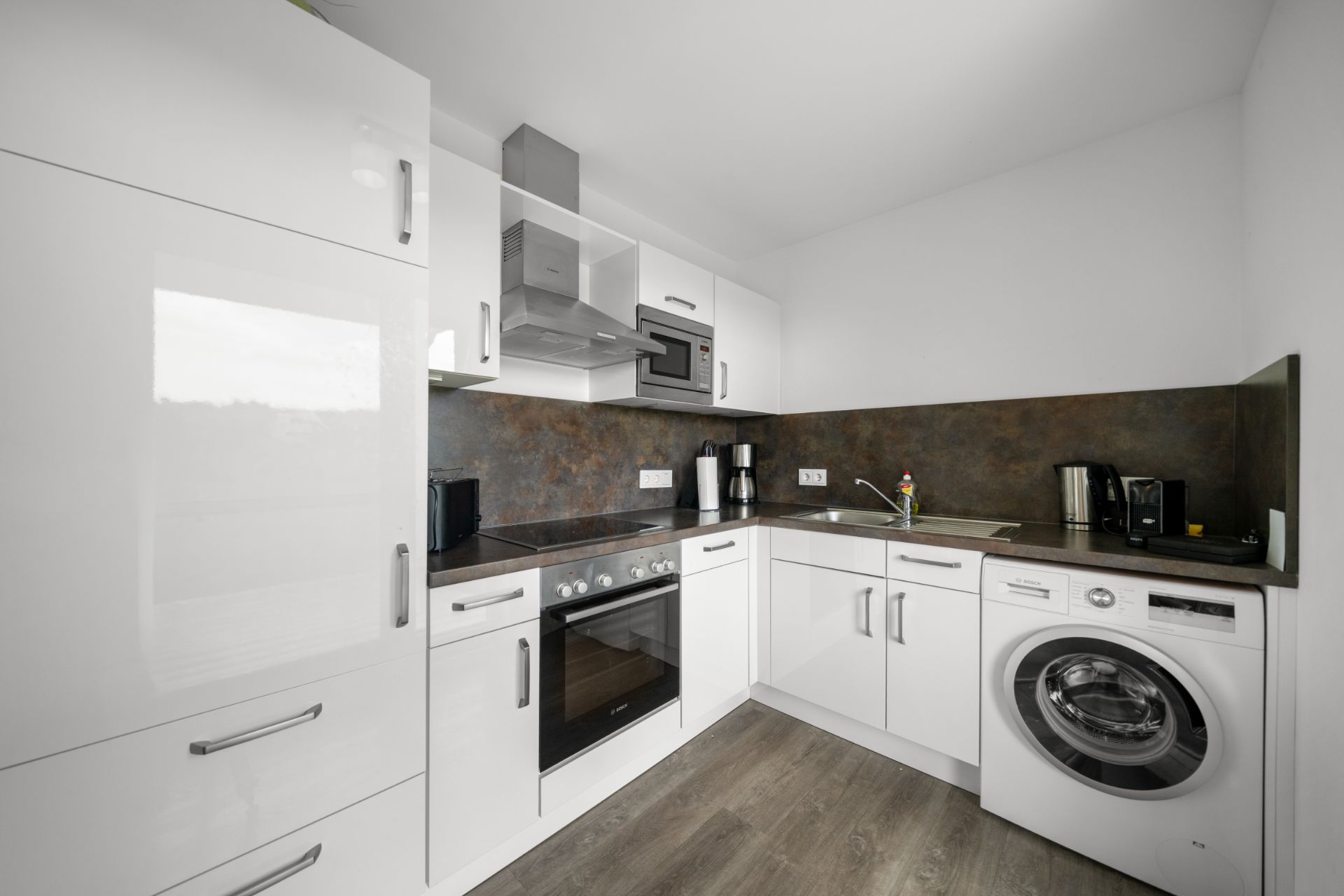 Moderne Einbauküche mit weißen Schränken, Backofen, Waschmaschine und Küchengeräten.