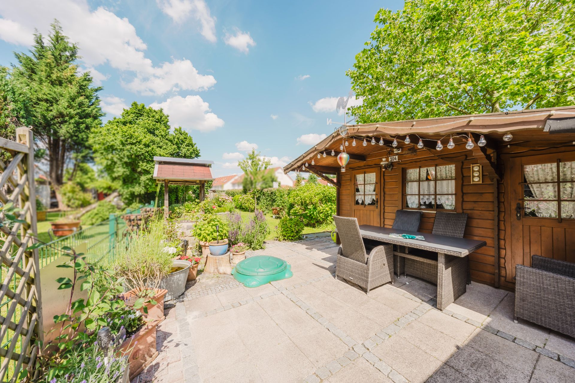 Gepflasterte Terrasse mit Holzhütte, Gartentisch, Stühlen und vielen Pflanzen im Garten.