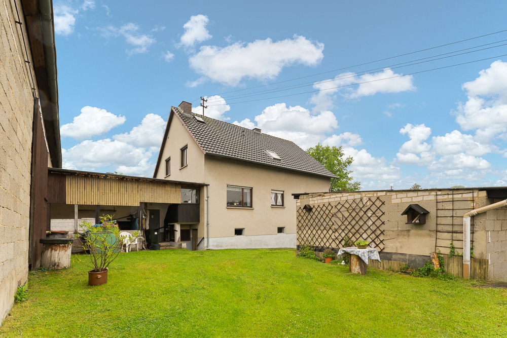 Haus mit Satteldach, kleinem Garten und angrenzenden Nebengebäuden bei blauem Himmel.