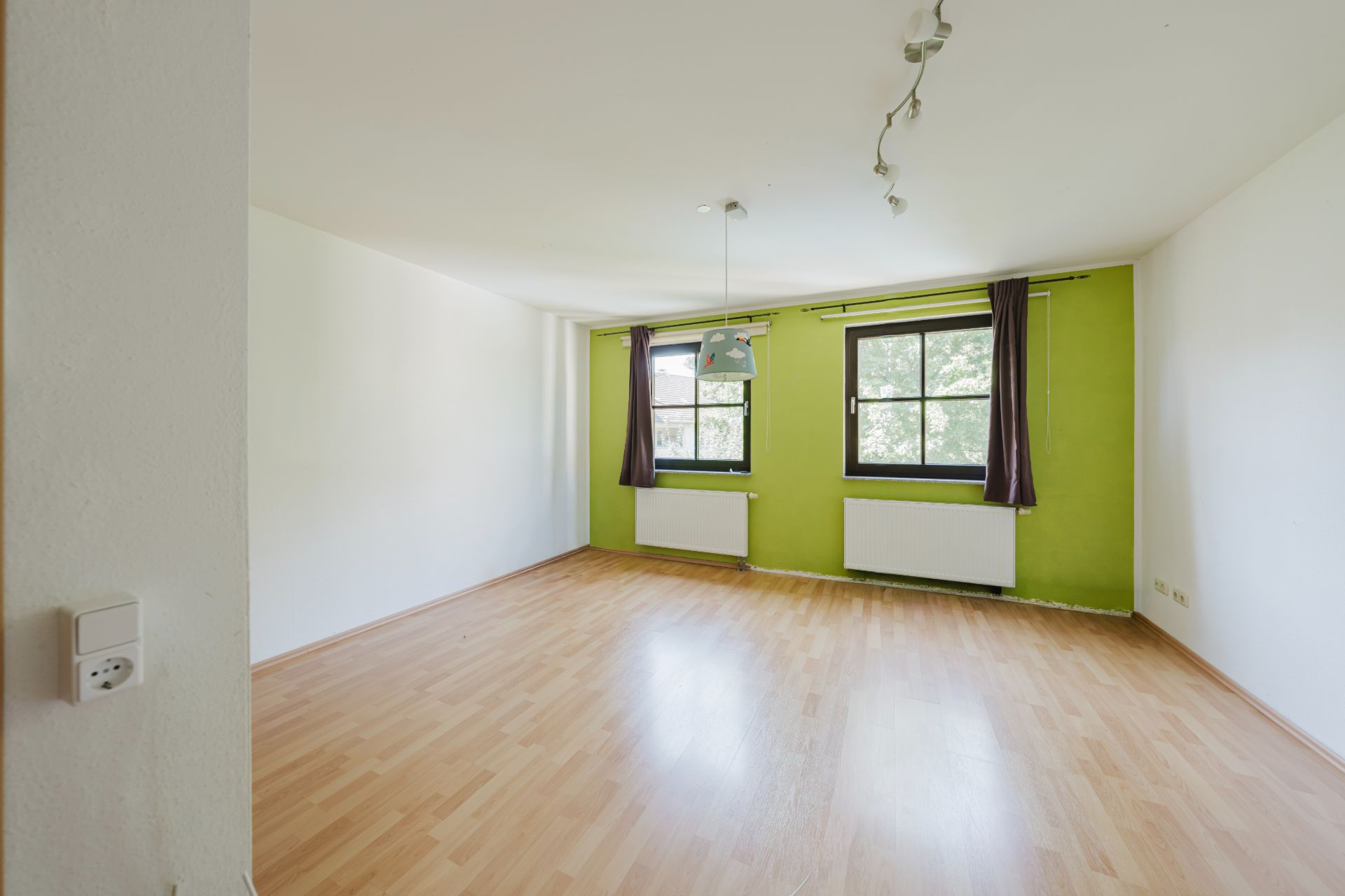 Leeres Zimmer mit hellem Laminatboden und zwei Fenstern vor grüner Wand, weiße Heizkörper darunter.
