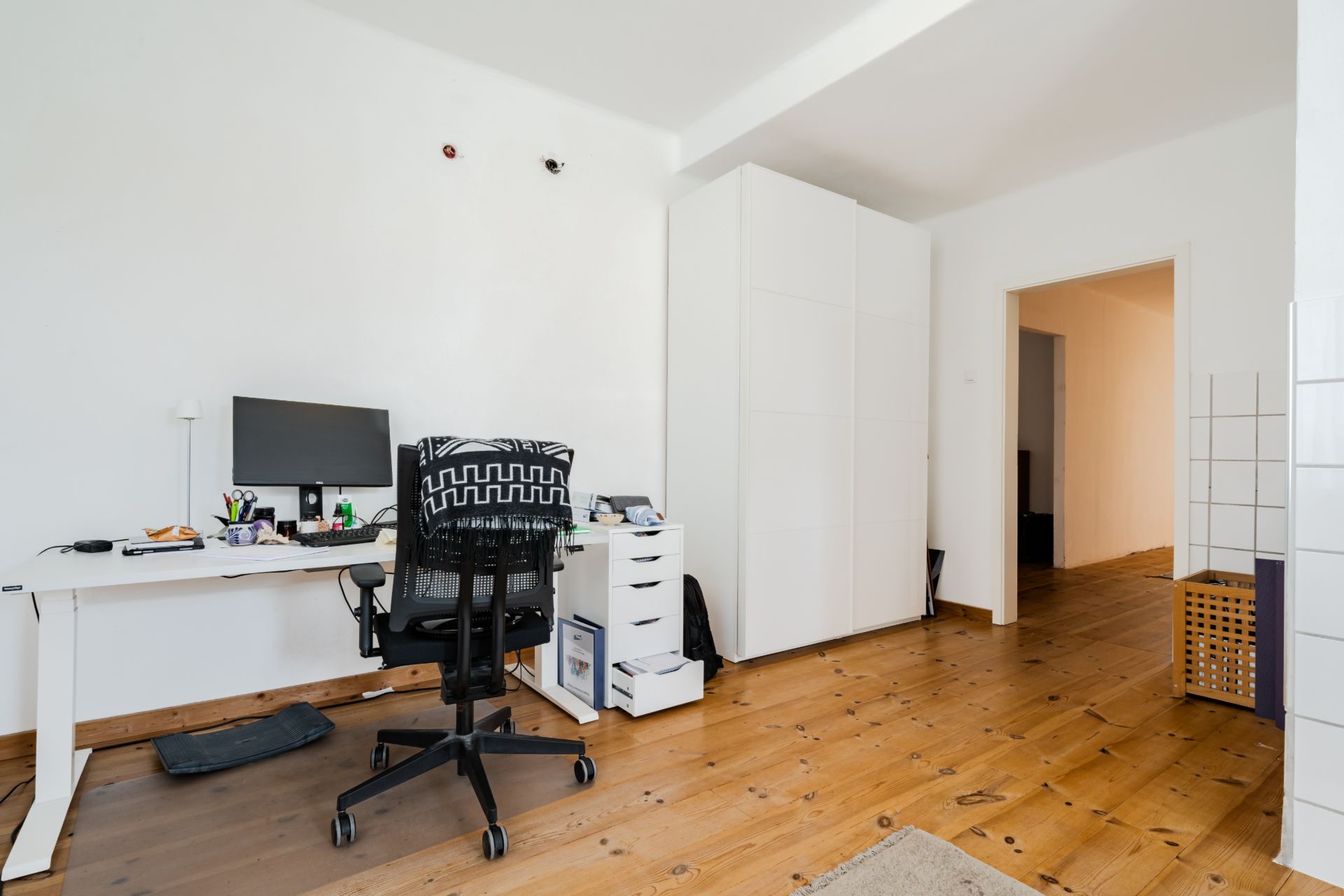 Helles Zimmer mit Schreibtisch, Bürostuhl, Computer und weißem Kleiderschrank auf Holzboden.