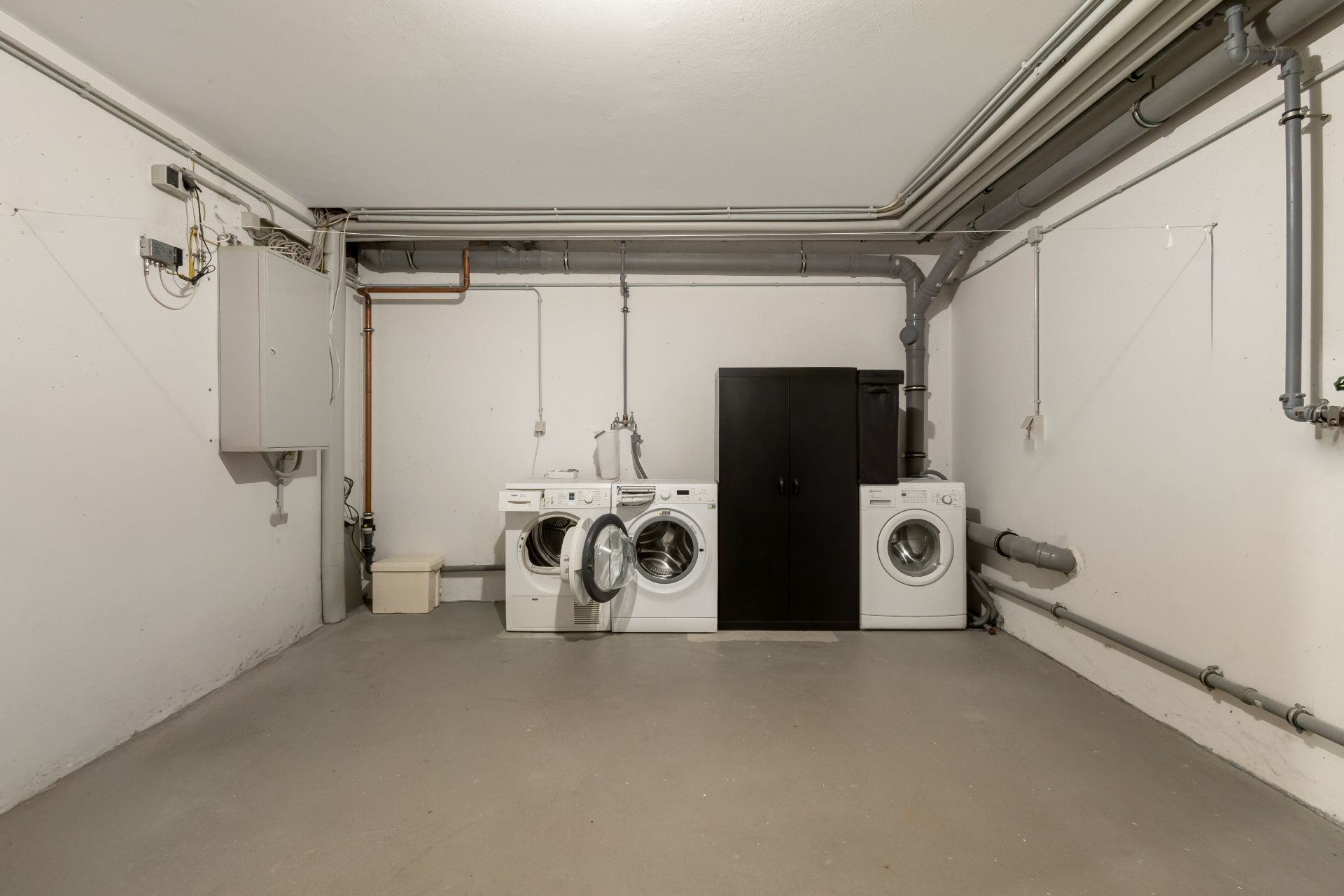 Waschkeller mit zwei Waschmaschinen und einem schwarzen Schrank an der Wand.
