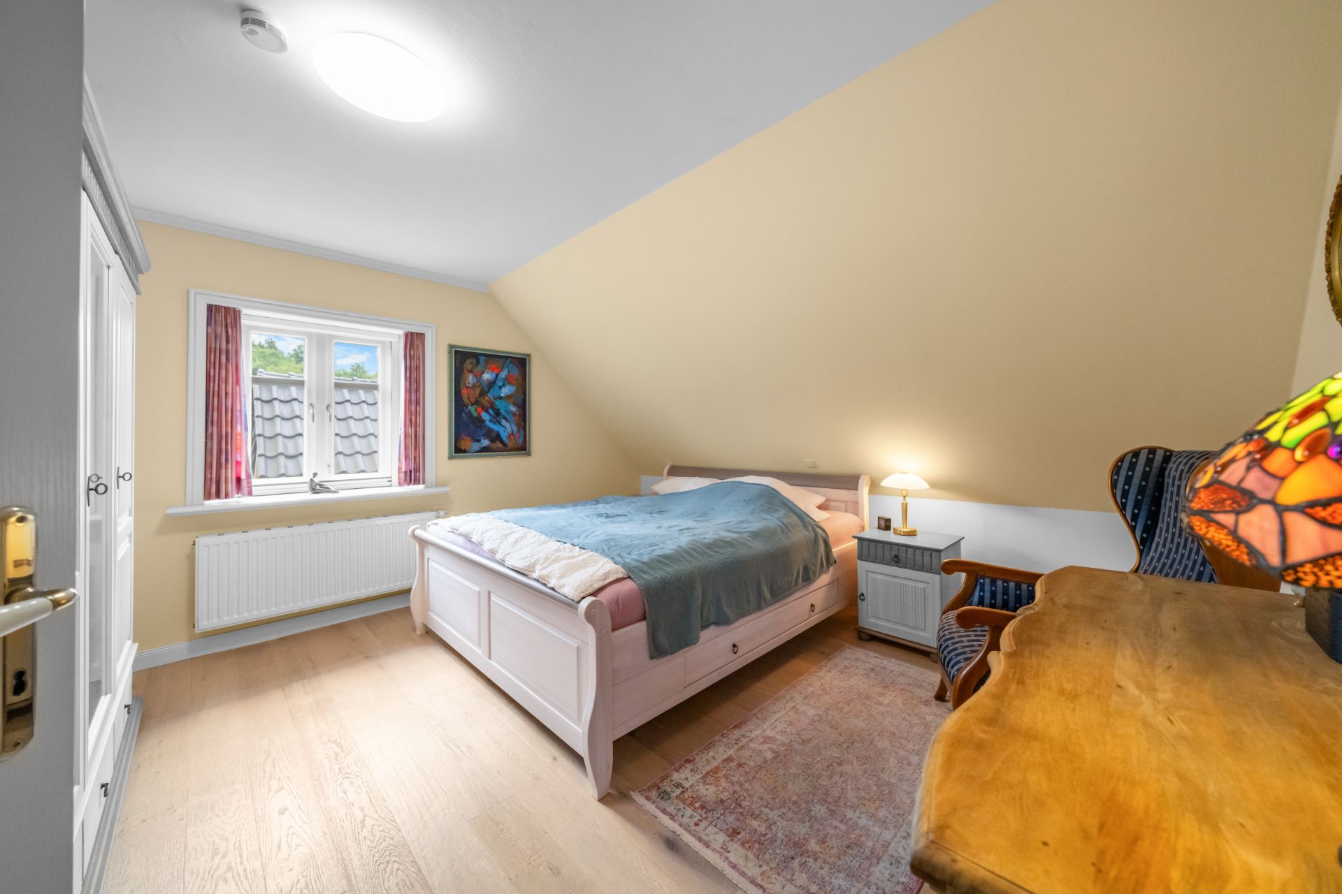 Schlafzimmer mit Doppelbett, Nachttisch, Stuhl, Schreibtisch und Fenster mit Vorhängen.