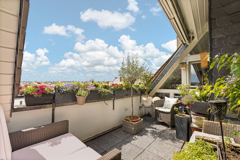Balkon mit Sitzmöbeln, vielen Blumen und Pflanzen, Blick auf die Stadt unter blauem Himmel.