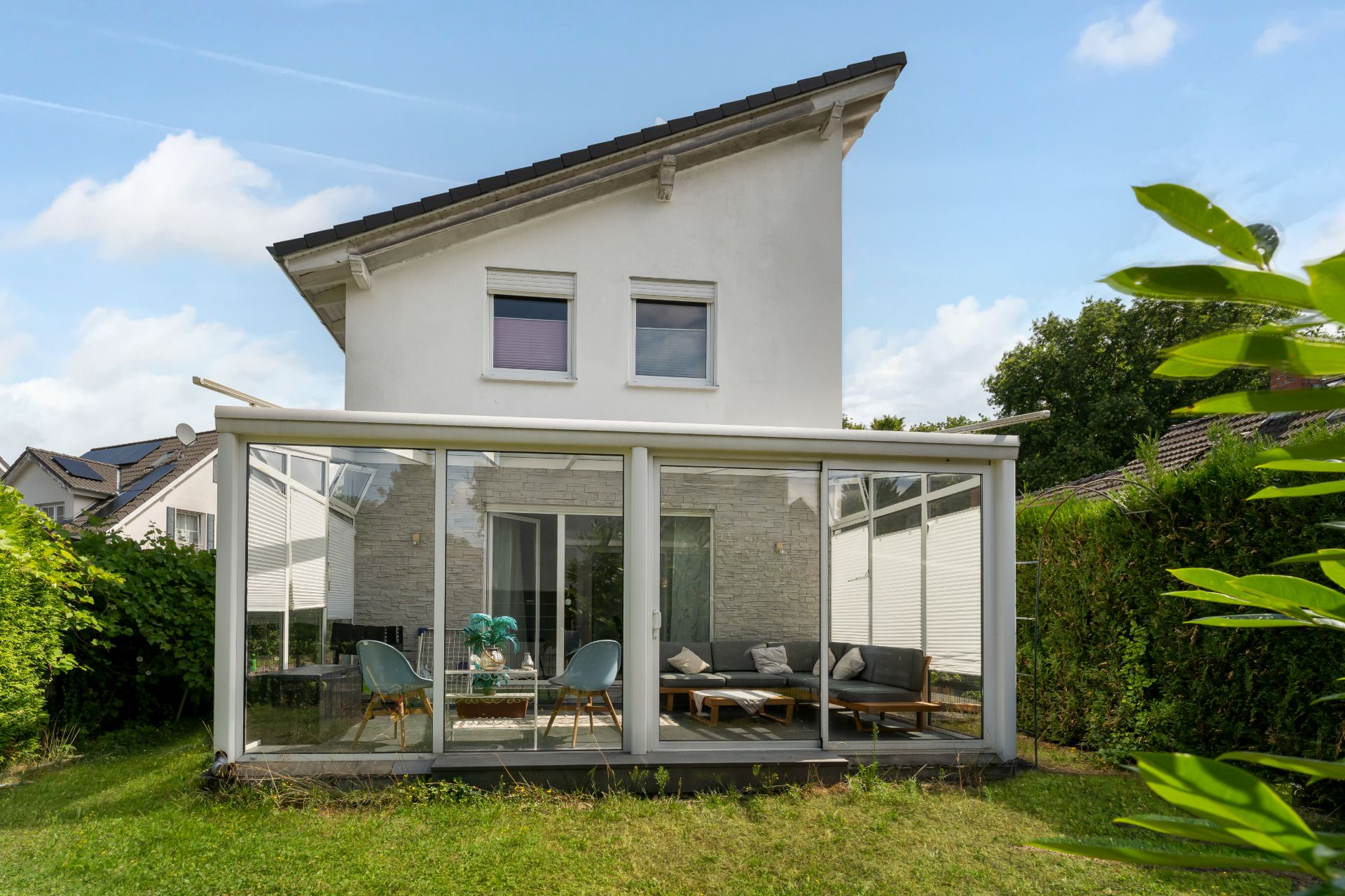 Zweistöckiges Haus mit Wintergarten und Gartenmöbeln, umgeben von grünem Rasen.