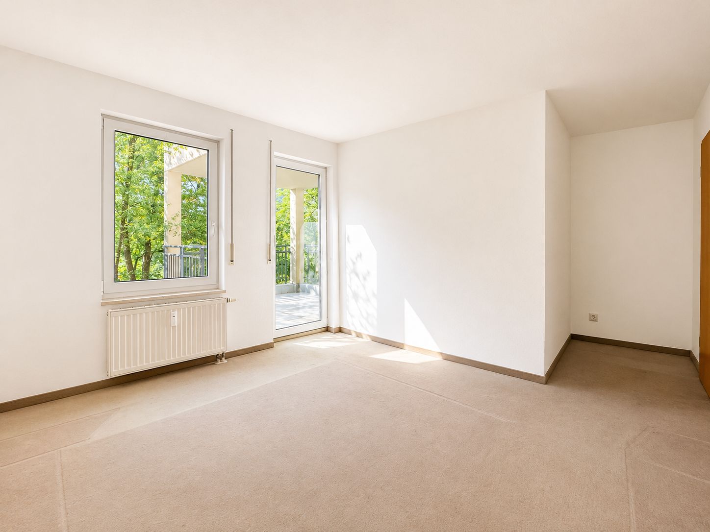 Helles, leeres Zimmer mit Teppichboden, Fenster und Tür zu einem Balkon.